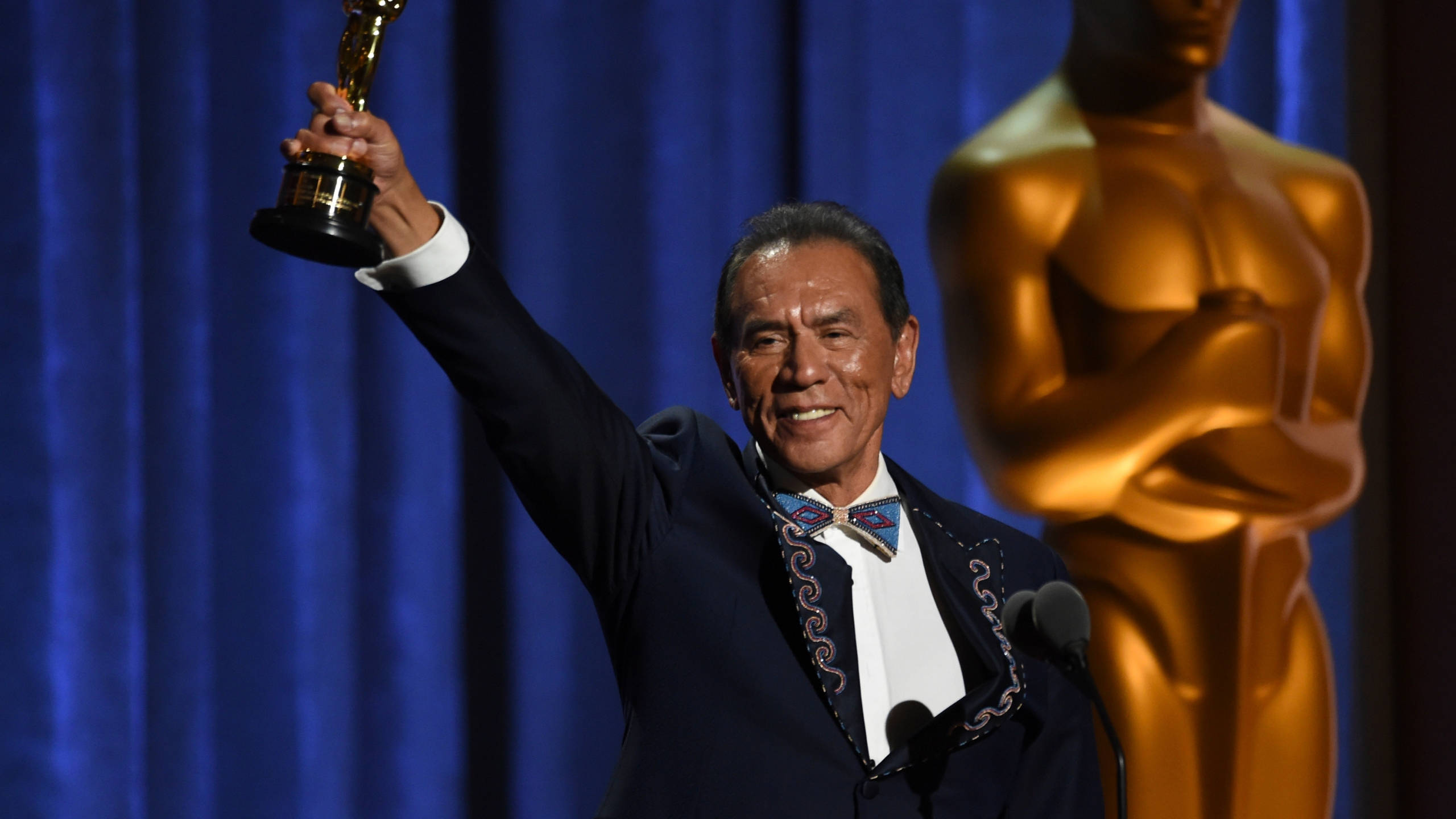 Wes Studi 領取奧斯卡獎 壁紙 — 一名身穿深色西裝、繫著圖案領結的男子，在藍色帷幕舞台上得意地高舉奧斯卡獎座，旁邊有一座金色奧斯卡雕像。