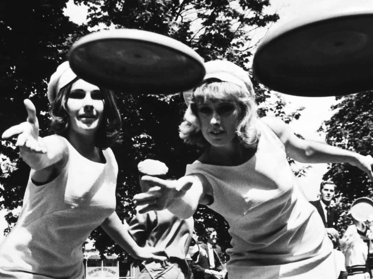Wham-O 玩具最佳 Ultimate Frisbee 背景 壁紙 — 兩位穿著復古服裝的女性在明亮的陽光下，以樹木為背景，在戶外玩飛盤。