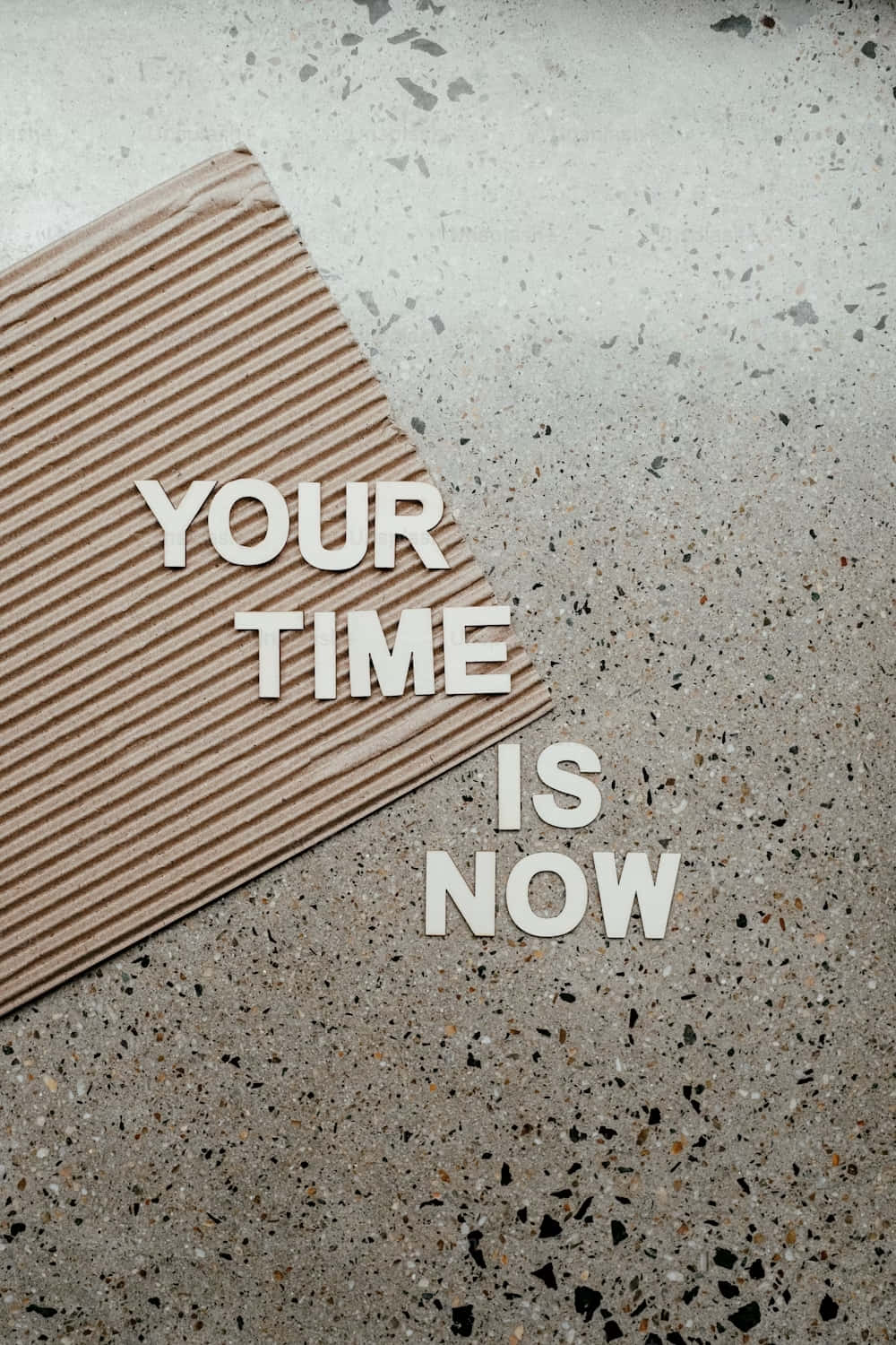  壁紙 — 白色3D字母拼出「YOUR TIME IS NOW」，放在一塊瓦通紙板上，置於斑點混凝土表面之上。