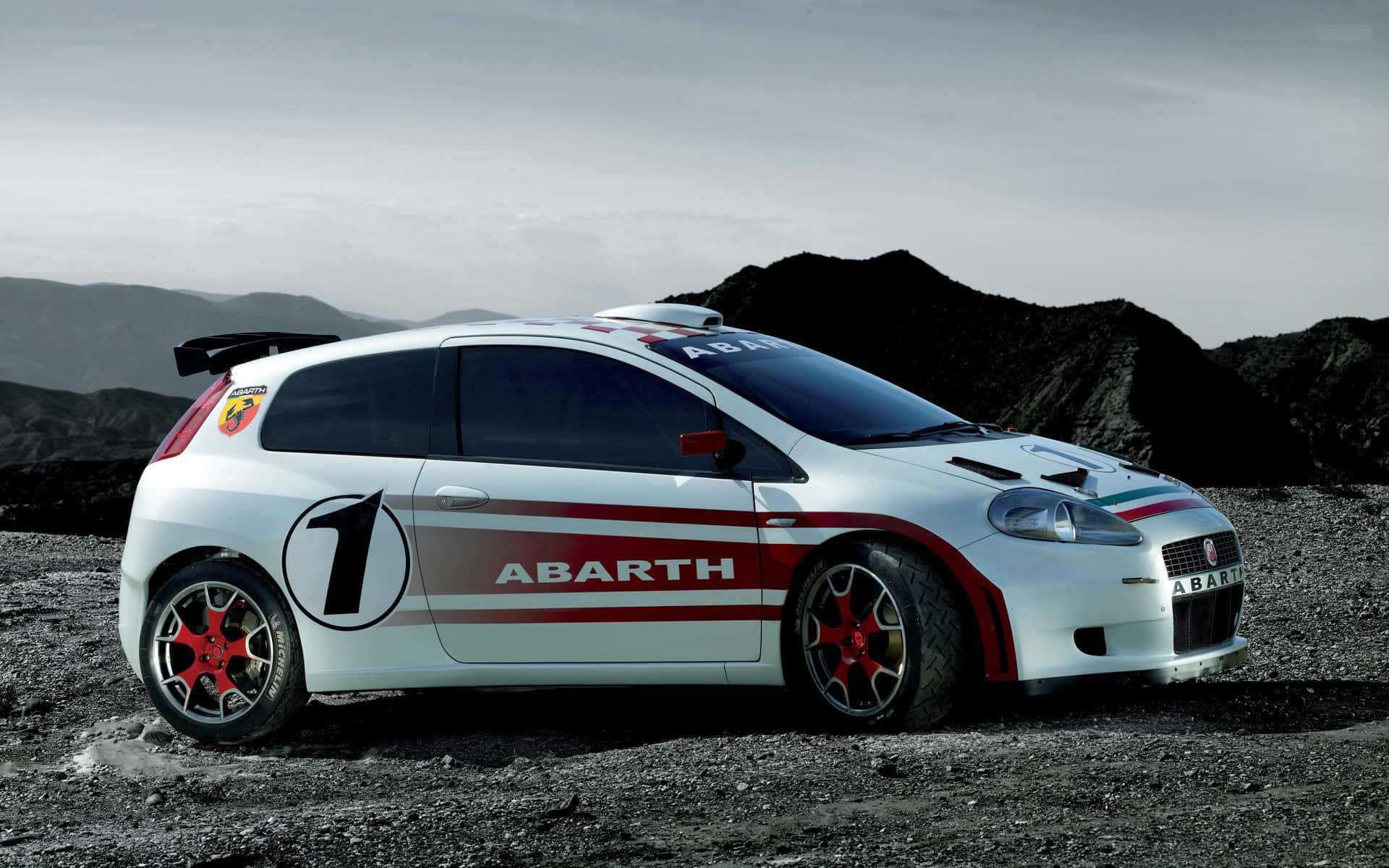 Fiat Grande Punto 桌布 壁紙 — 一輛帶有紅色裝飾和數字一的白色Abarth拉力賽車，在陰沉天空下，以黑暗火山丘陵為背景，停在碎石地上。