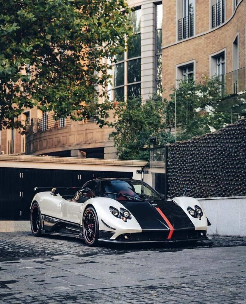 Pagani Zonda C12 Roadster 桌布 壁紙 — 一輛帶有紅色點綴的白色和黑色Pagani跑車停泊在鵝卵石街道上，旁邊是現代建築和綠樹。