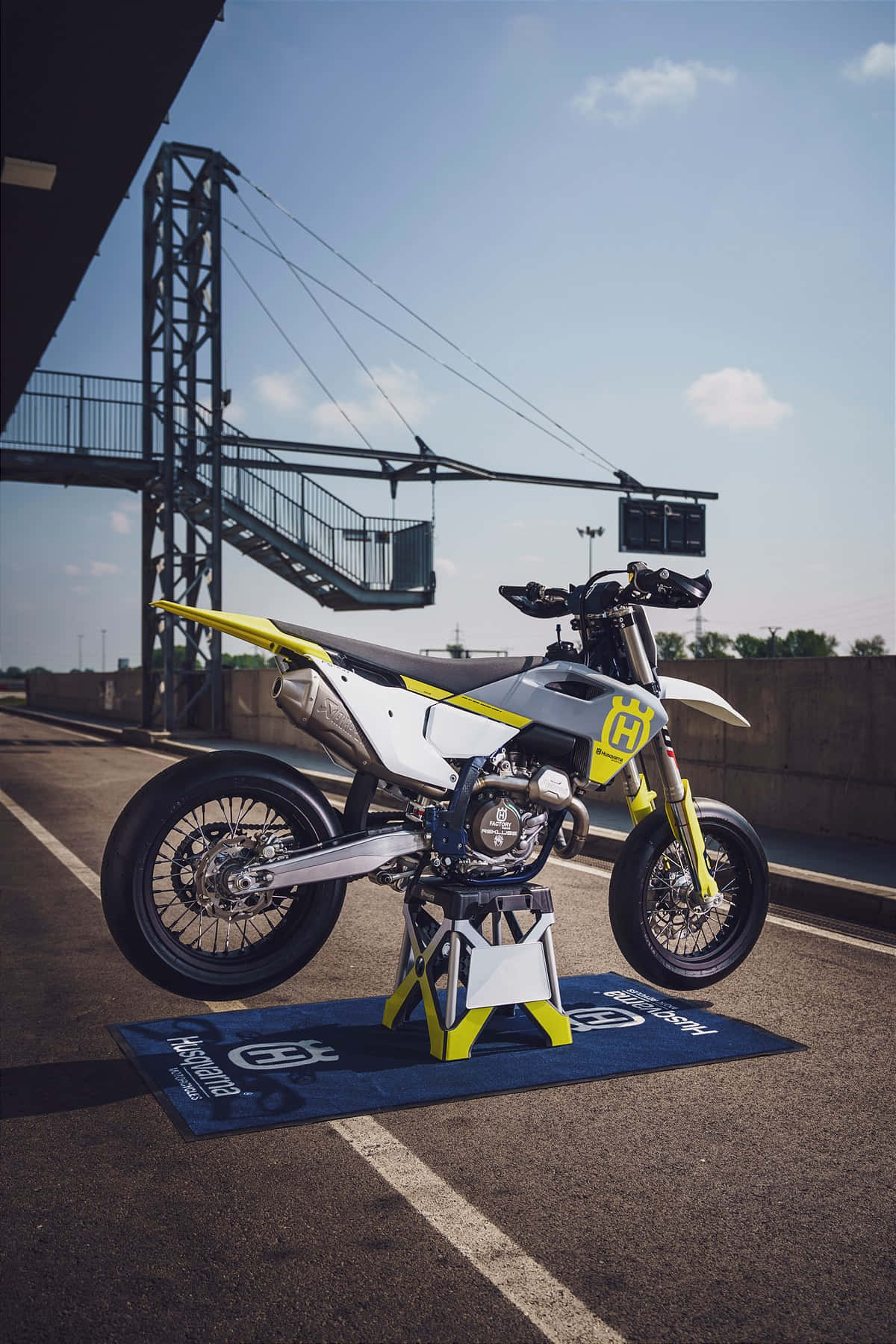  壁紙 — 一架白色和黃色的 Husqvarna 越野電單車，在賽車場的藍色品牌墊上，於晴朗的藍天下展示。