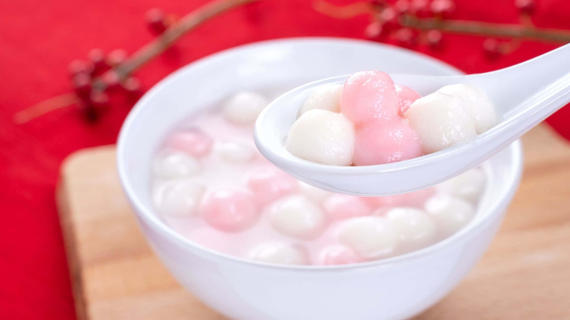  壁紙 — 一隻白色陶瓷湯匙從一碗甜奶湯中舀起粉紅色和白色的糯米糰，背景是鮮紅色的櫻花枝。