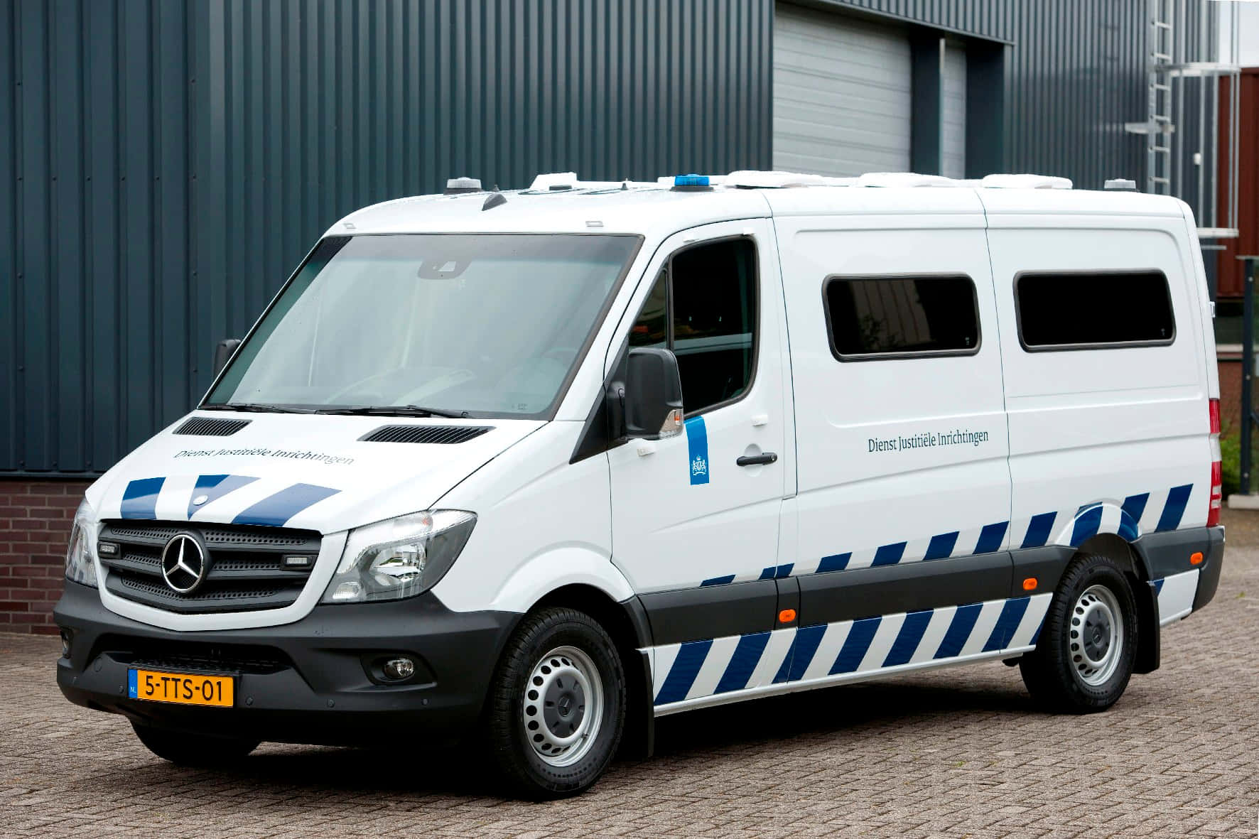 Mercedes Benz Sprinter 桌布 壁紙 — 一輛白色荷蘭警車，帶有藍色和黑色斜紋，停在深灰色工業建築前的磚砌路面上。