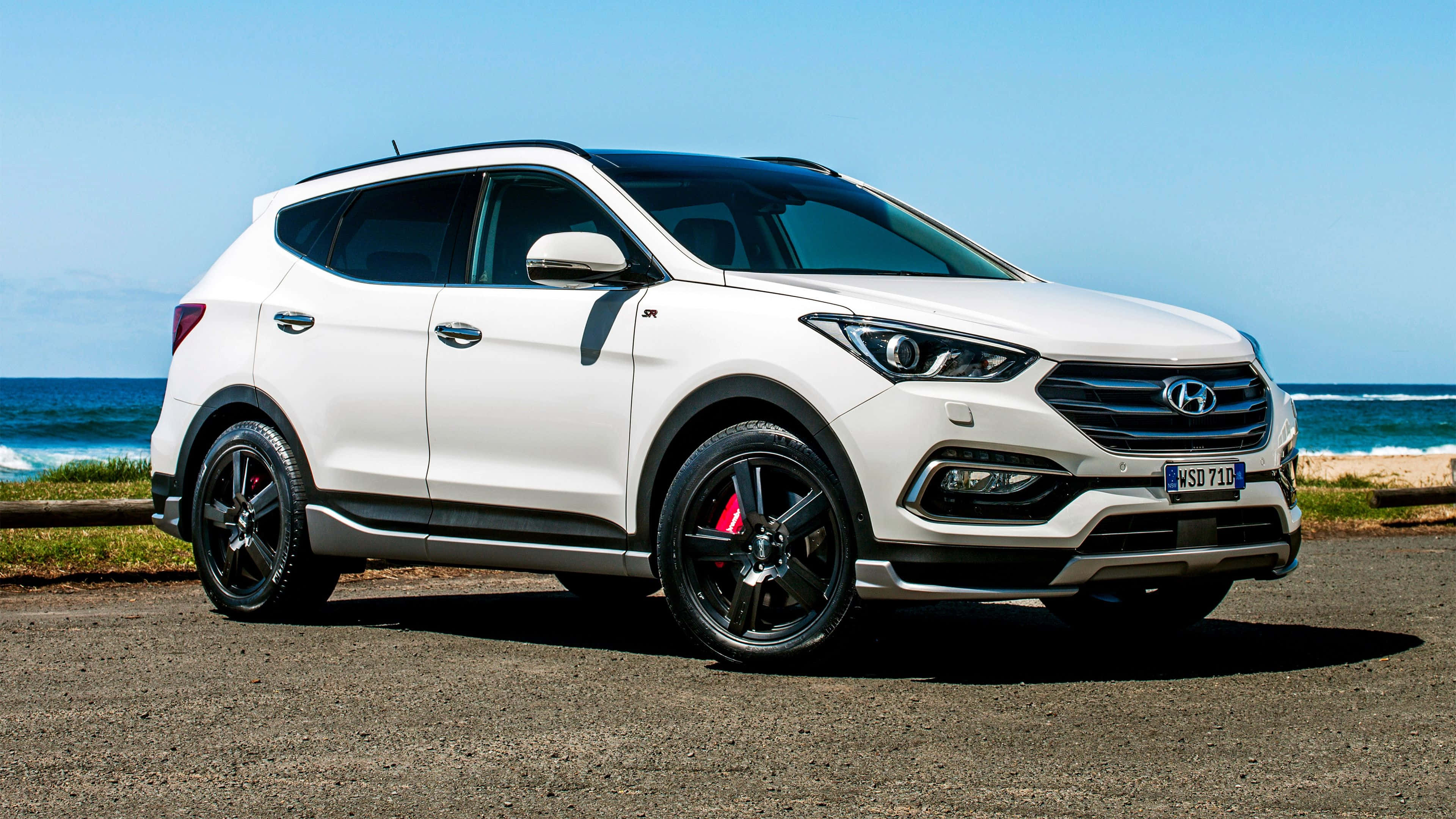 Hyundai Santa Fe 桌布 壁紙 — 一輛白色現代Santa Fe SUV，配備黑色輪圈和紅色制動卡鉗，停泊在海洋旁邊的碎石路上，天空清澈湛藍。