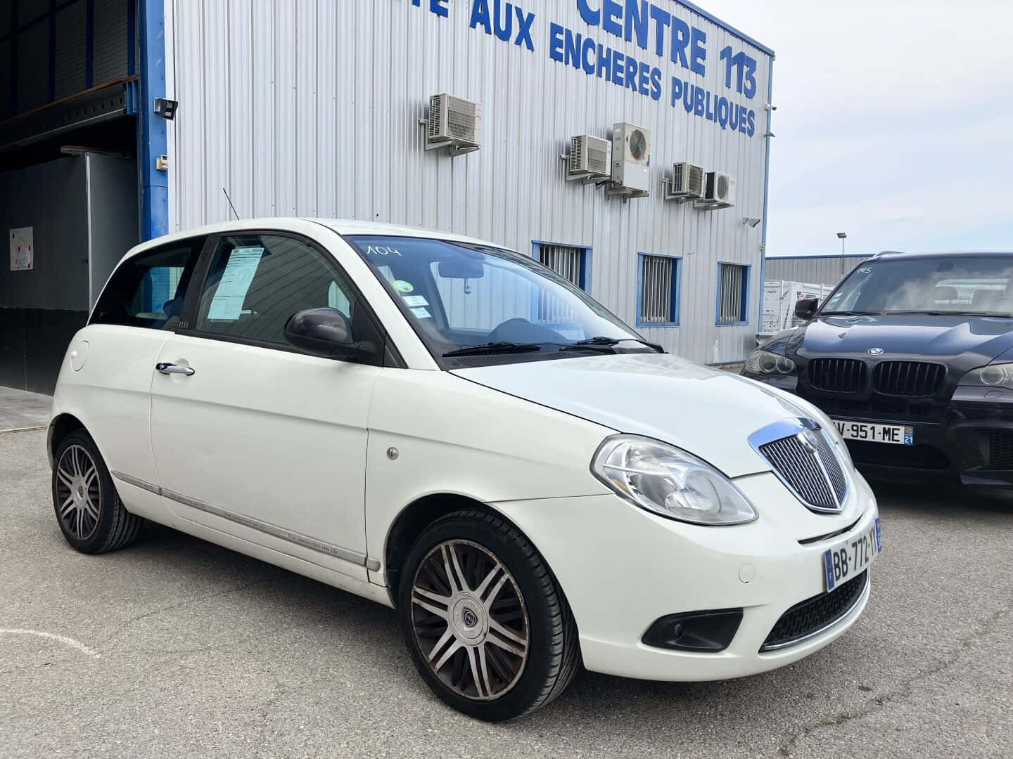  壁紙 — 一輛白色 Lancia Ypsilon 停泊在公共拍賣中心外的戶外，旁邊有一輛黑色 BMW。