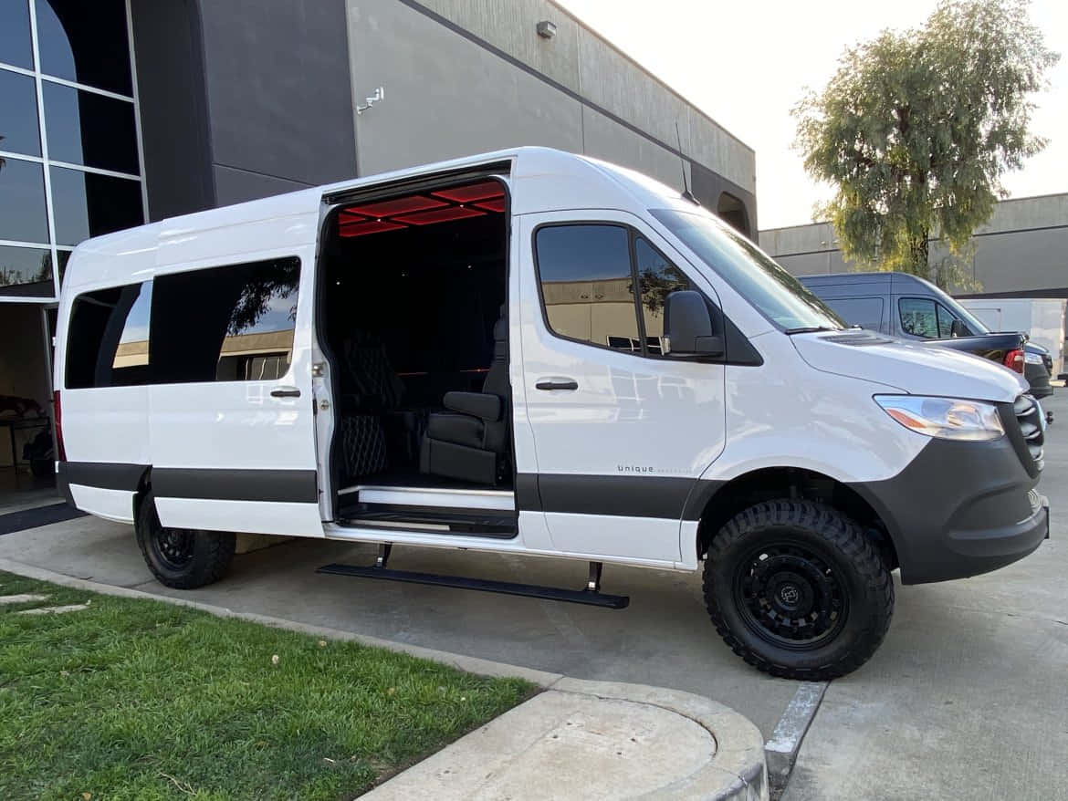 Mercedes Benz Sprinter 桌布 壁紙 — 一輛白色豪華客貨車，帶有黑色飾邊和堅固輪胎，停在一棟現代建築外，側門打開，露出紅色內部照明。