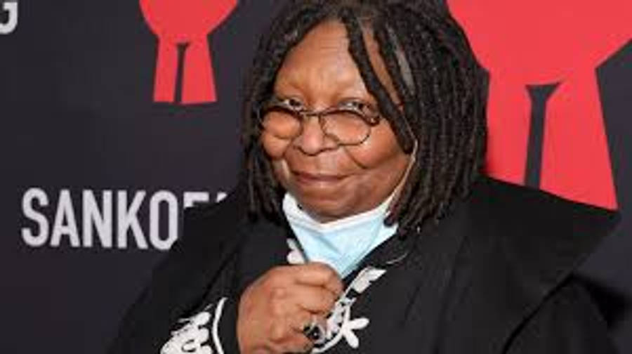 Whoopi Goldberg 背景為 Sankofa.org 布幔 壁紙 — 一位戴眼鏡和辮子頭、面帶微笑的女性在帶有 'SANKOF' 品牌的紅地毯活動中調整她的口罩。