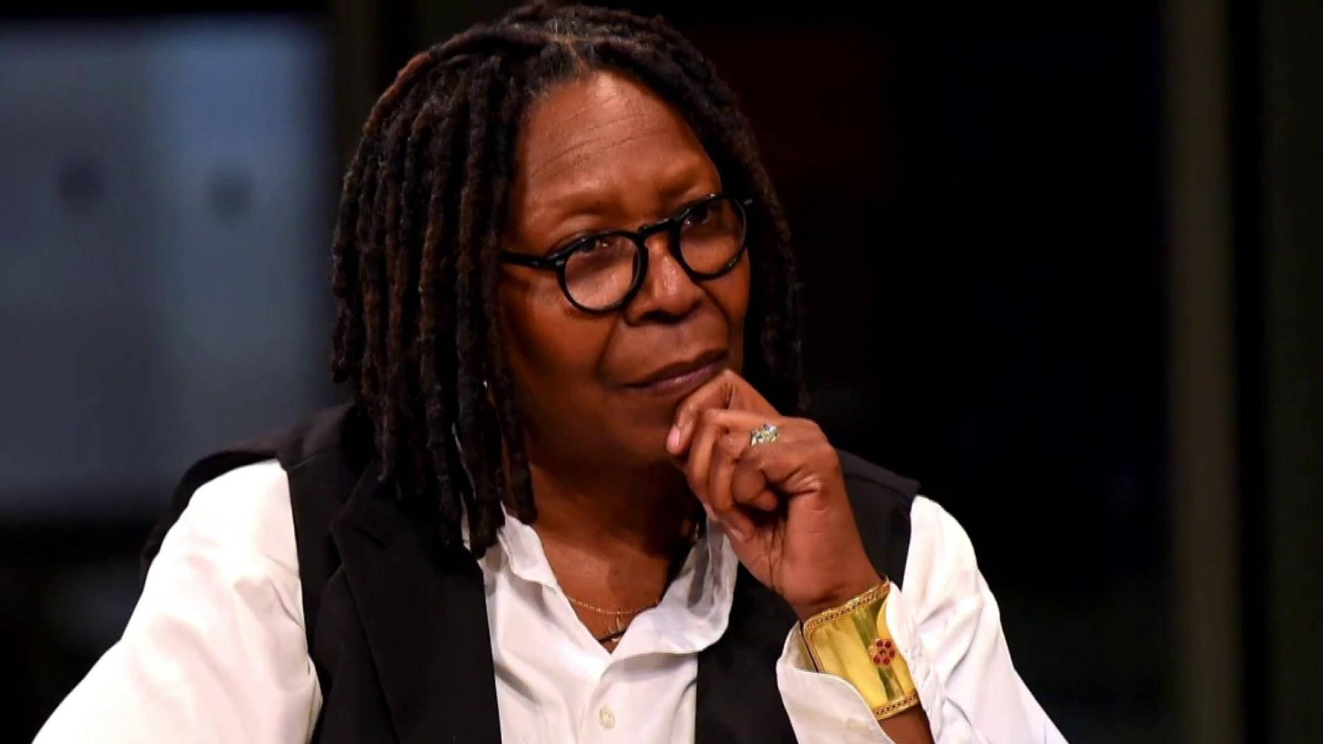Whoopi Goldberg 談話節目 壁紙 — 一位戴著辮子頭和眼鏡的深思女性，用手托著下巴，身穿白色襯衫和黑色背心。