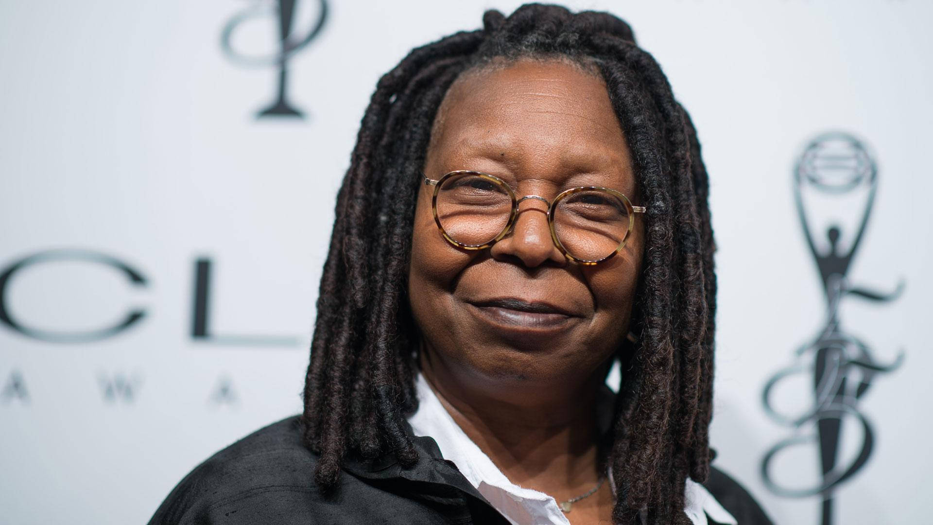 Whoopi Goldberg 紅毯頒獎時的風采 壁紙 — 一位微笑的女士，留著長辮子和戴圓框眼鏡，在頒獎活動中於品牌背景前擺姿勢。