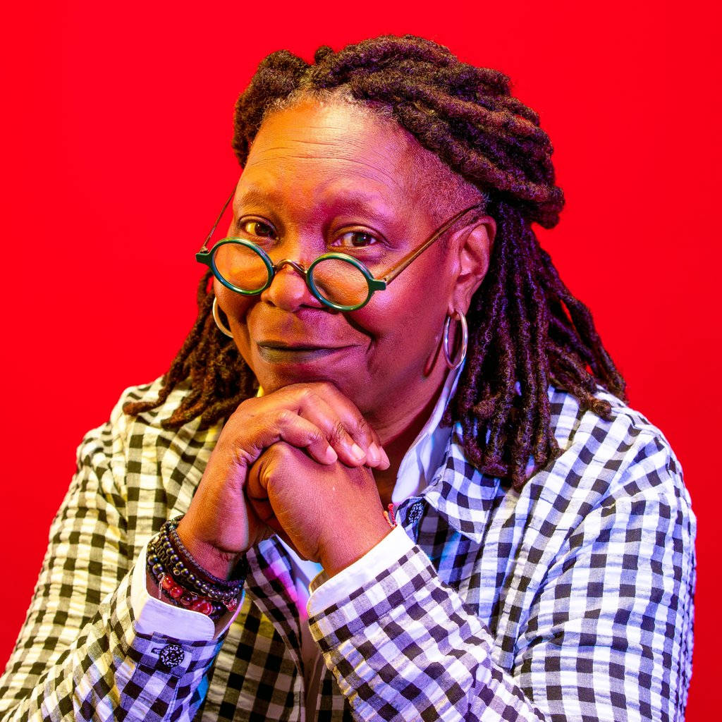 Whoopi Goldberg 紅色背景 壁紙 — 一位留著辮子、戴圓框眼鏡的女性，在鮮紅色背景前，雙手交疊托著下巴，溫暖地微笑。