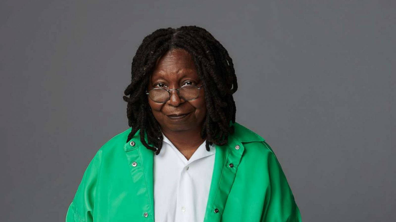 Whoopi Goldberg 身穿鮮豔霓虹綠夾克展現時尚 壁紙 — 一位戴著眼鏡和辮子頭的年長黑人女性，穿著亮綠色外套，在灰色工作室背景下的溫暖肖像。