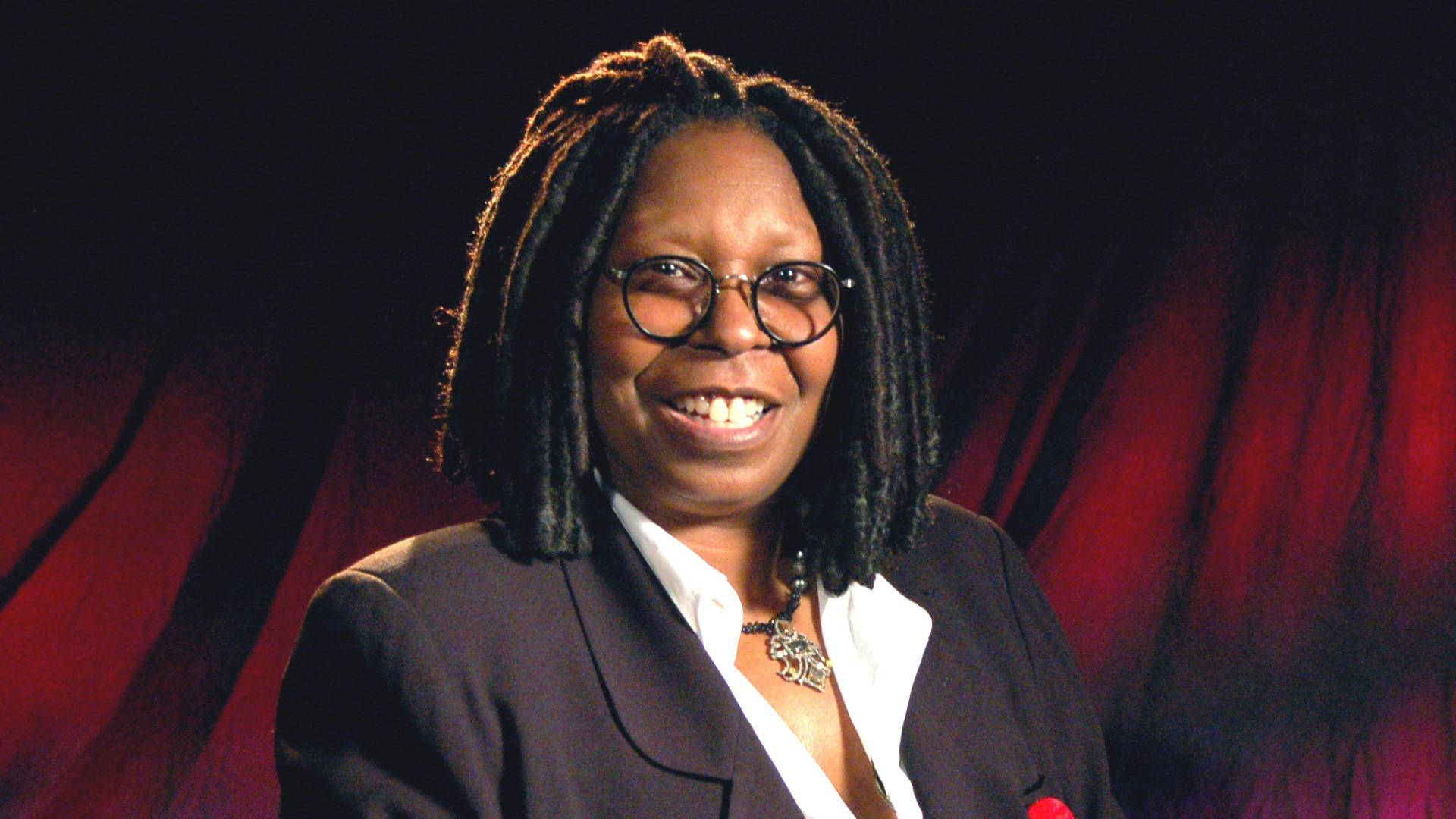 Whoopi Goldberg 正式服裝 壁紙 — 一位戴眼鏡和辮子頭的女性微笑著，在紅色窗簾背景下穿著深色西裝外套內搭白襯衫。