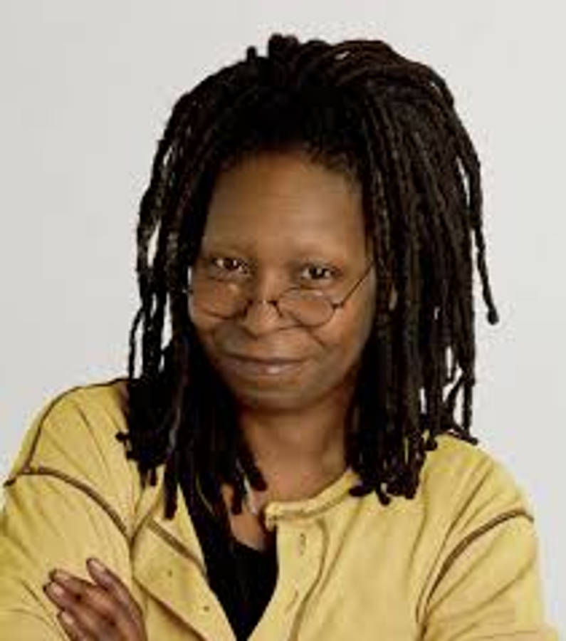 Whoopi Goldberg 黃色襯衫 壁紙 — 一位擁有深色辮子和眼鏡的女性，在純色背景前穿著黃色外套，溫柔地微笑。