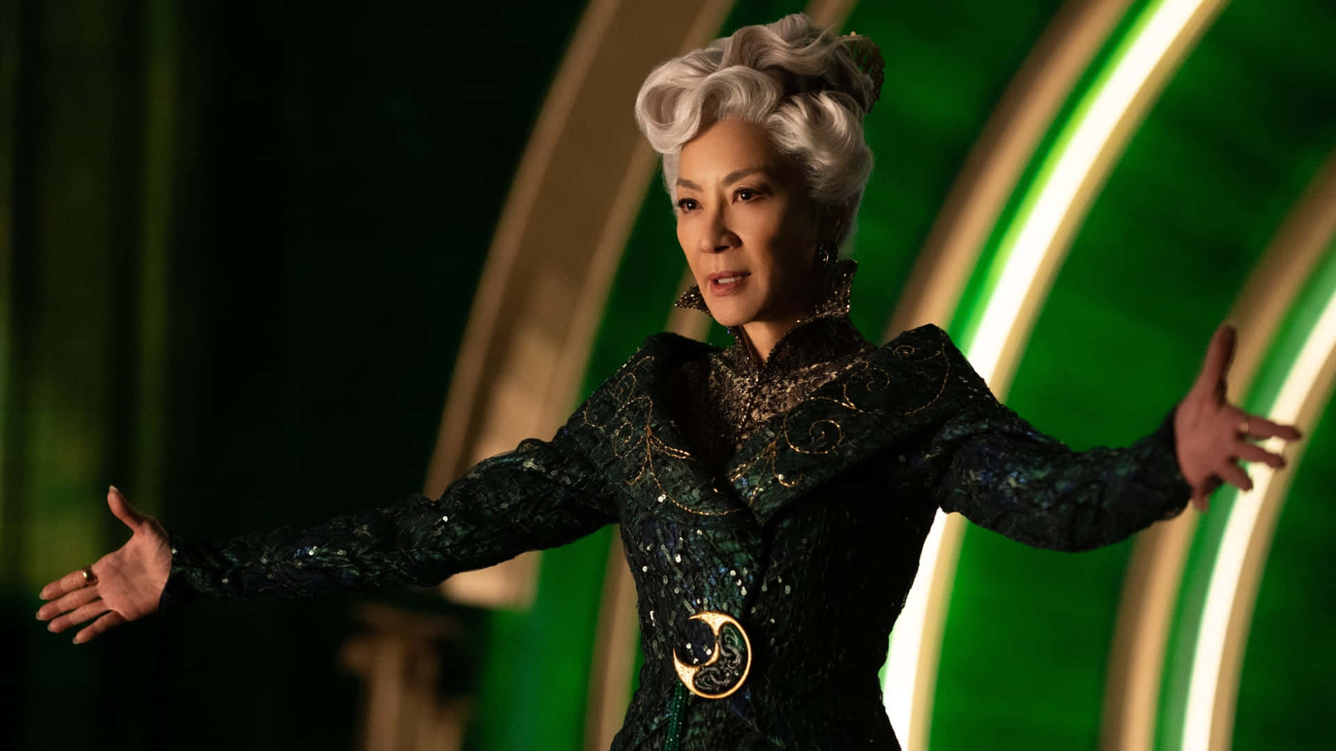 《Wicked》電影中的艾爾法巴演出 壁紙 — 一位高貴的白髮捲曲女性，身穿華麗的深綠色亮片禮服，飾有金色點綴和新月形腰帶扣，雙臂伸展在發光的綠色拱門前。