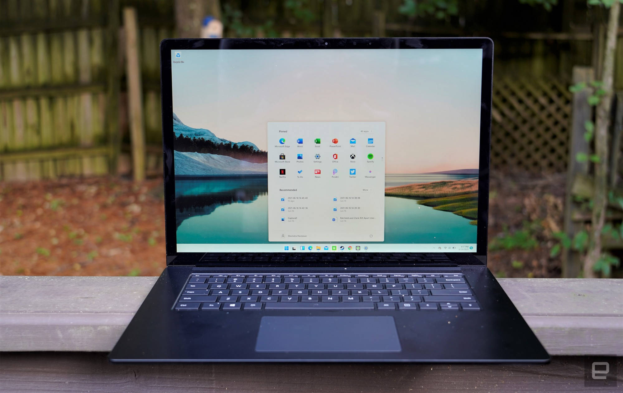 Windows 11 筆電陽台 壁紙 — 一部黑色手提電腦打開放在木長凳上，顯示寧靜的山湖壁紙，上面覆蓋著應用程式圖標。