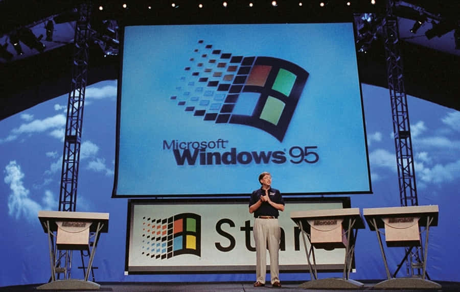 Windows 95 – 升級您的作業系統 壁紙 — 一名男子站在舞台上，巨型屏幕顯示著經典的Microsoft Windows 95標誌，背景是藍天。