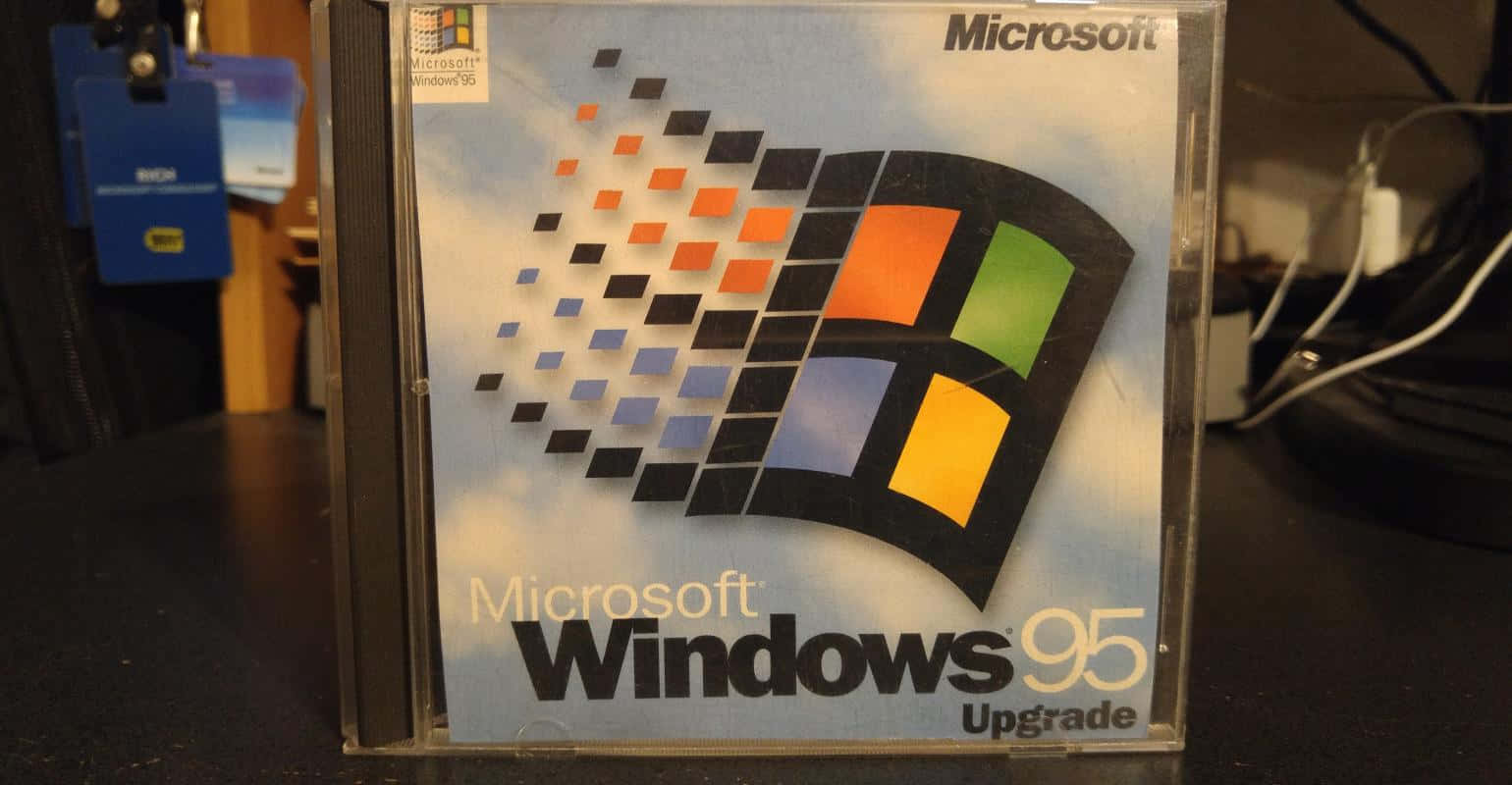 Windows 95 的經典發佈時刻 壁紙 — 一個復古的 Microsoft Windows 95 升級版 CD 盒放在深色桌子上，背景是模糊的科技背景。