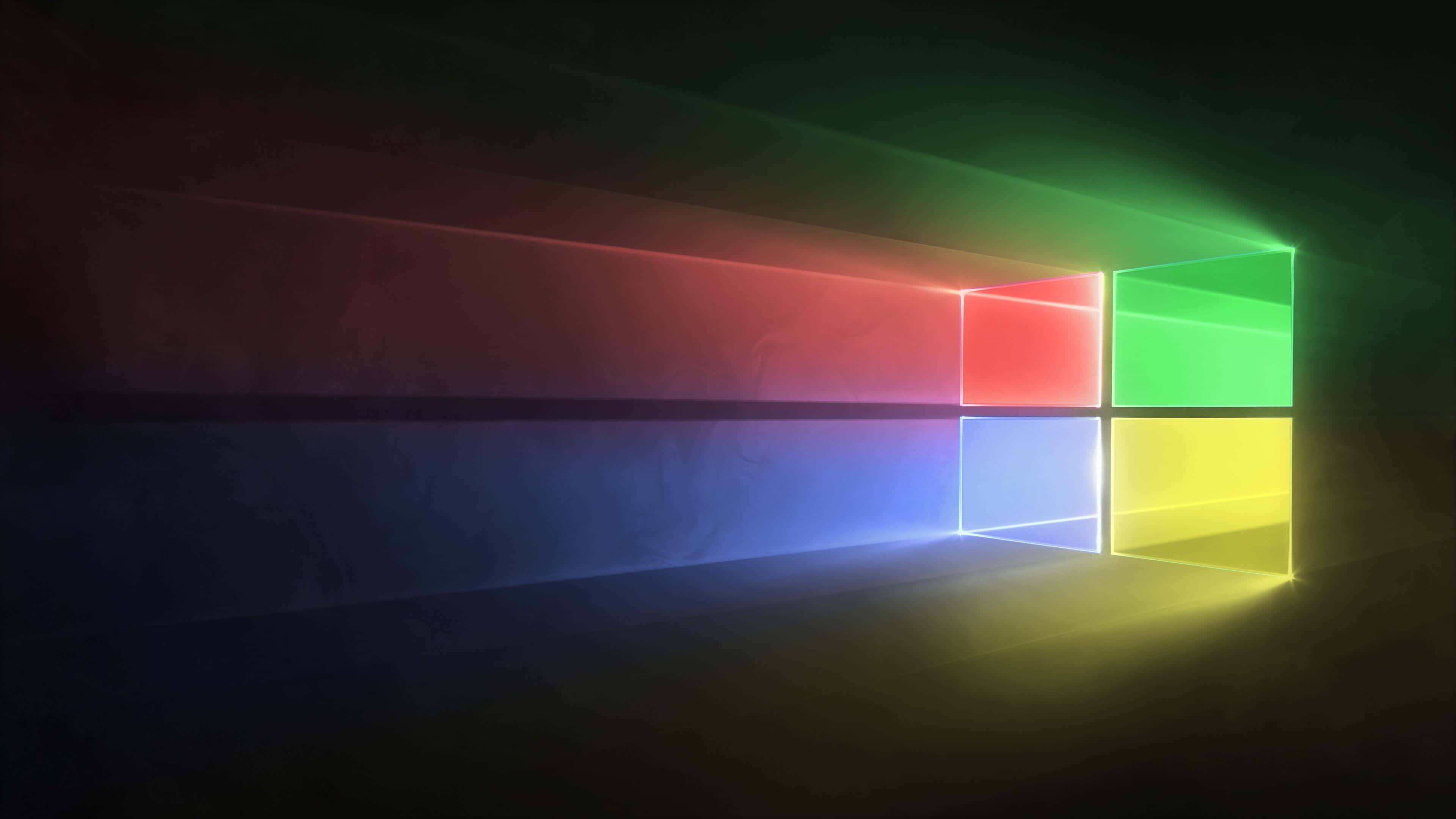 Windows 預設背景的近距離視角 壁紙 — 一個發光的Windows標誌在黑暗的房間中發出鮮豔的紅、綠、藍和黃色光束。