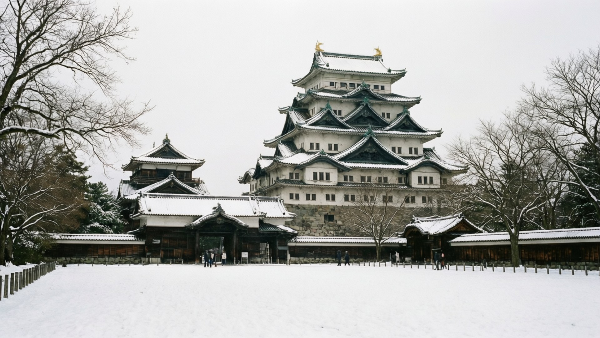 一座歷史悠久的日本城堡被白雪覆蓋，有光禿禿的樹木和遊客在附近行走。