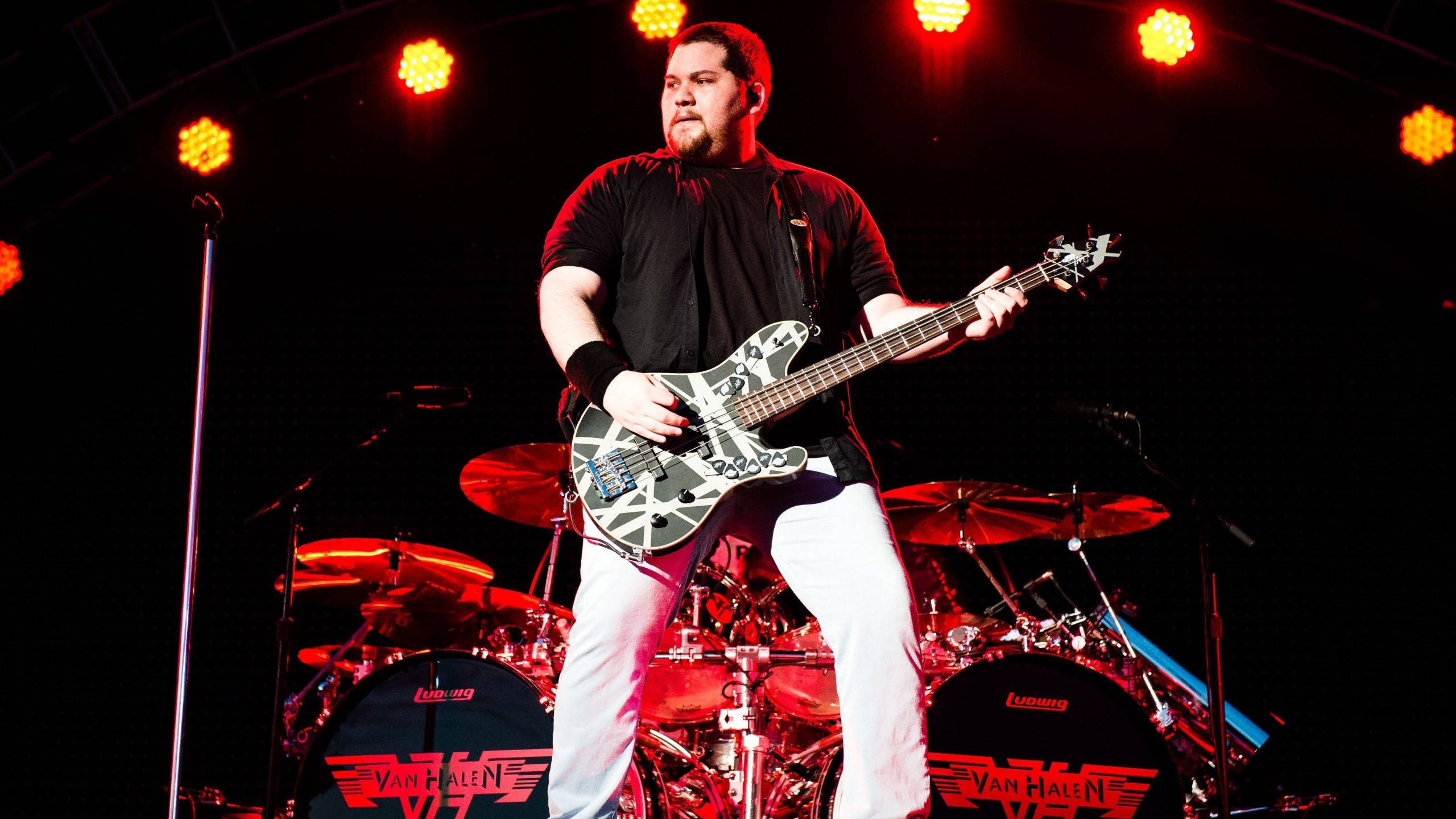 Wolfgang Van Halen 樂隊演奏 壁紙 — 一位留鬍鬚的結他手，身穿黑色襯衫和白色褲子，在紅色舞台燈光下彈奏黑白圖案結他，身後有爵士鼓。