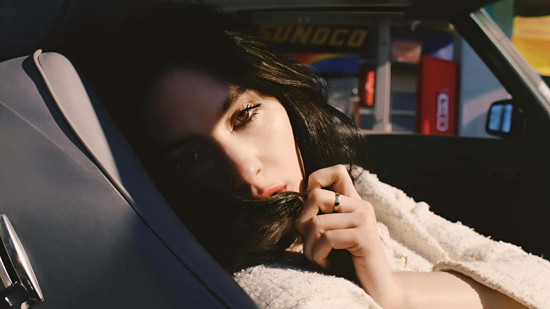  壁紙 — 一位深色頭髮的女性將頭靠在汽車座椅上，陽光照射在她的臉上，她帶著沉思的表情向前凝視。