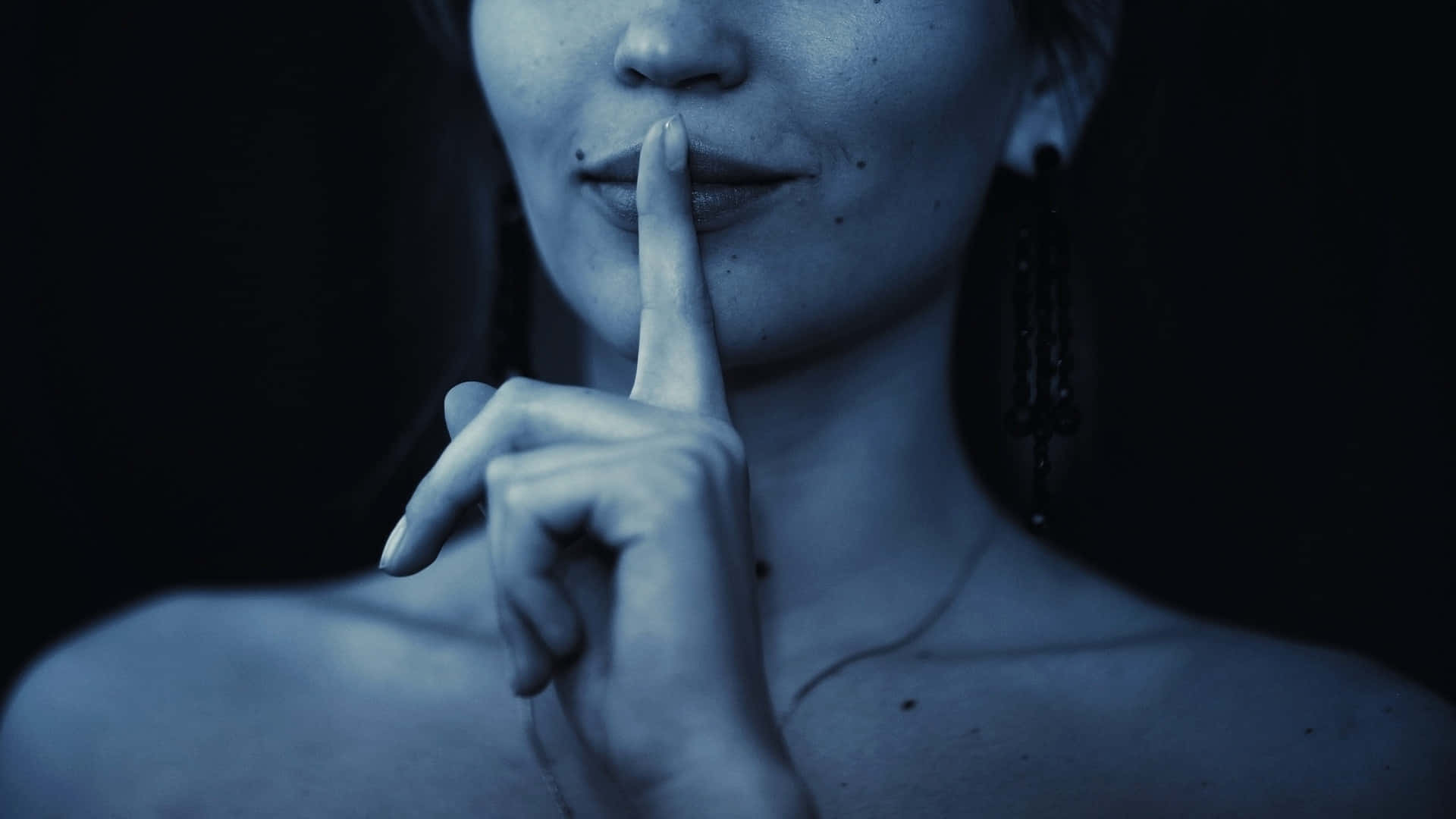  壁紙 — 一位身處冷藍色調的女性在黑暗背景前做出安靜的手勢，將手指放在嘴唇上。