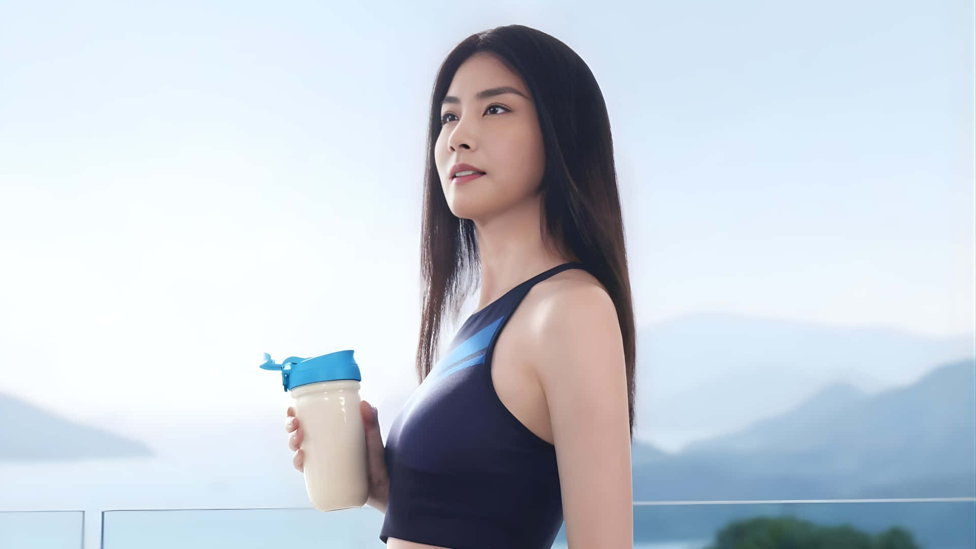  壁紙 — 一位穿著運動服的女性在陽台上手持蛋白奶昔，俯瞰著晴朗天空下的霧濛濛山脈。