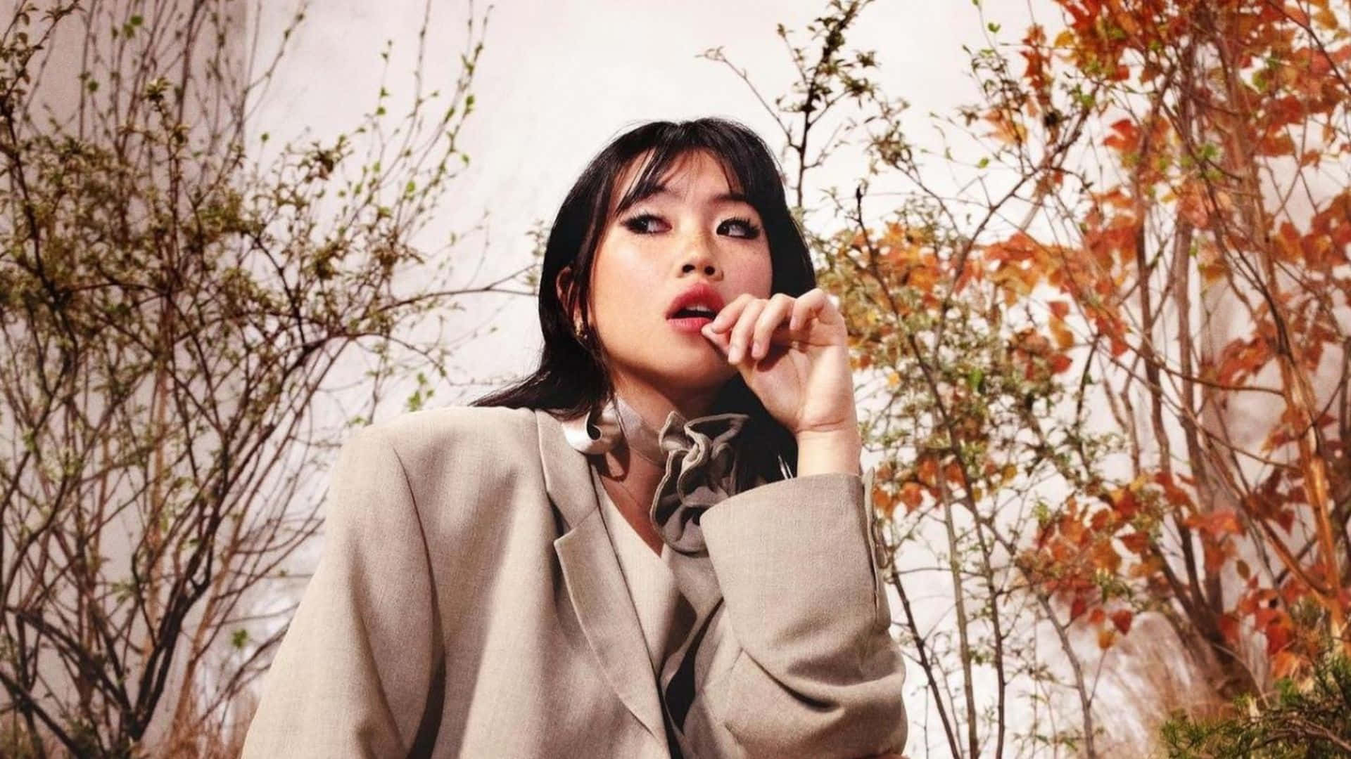  壁紙 — 一位身穿米色西裝外套的女性，在秋葉（橙色樹葉和光禿禿的樹枝）前若有所思地擺姿勢。