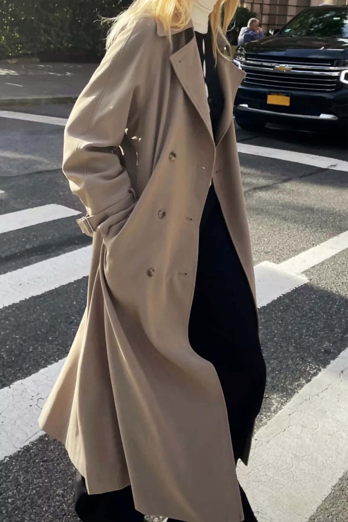  壁紙 — 一位身穿米色風衣的女性，在陽光明媚的城市街道上走過，街道上有白色斑馬線條紋，背景是一輛黑色SUV。
