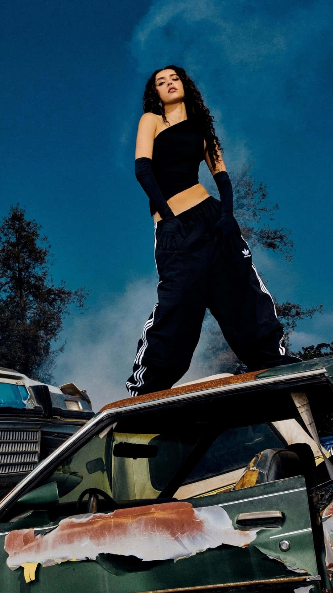  壁紙 — 一位身穿黑色運動服的女性自信地站在一輛生鏽的綠色汽車上，背景是湛藍的天空和縷縷白雲。
