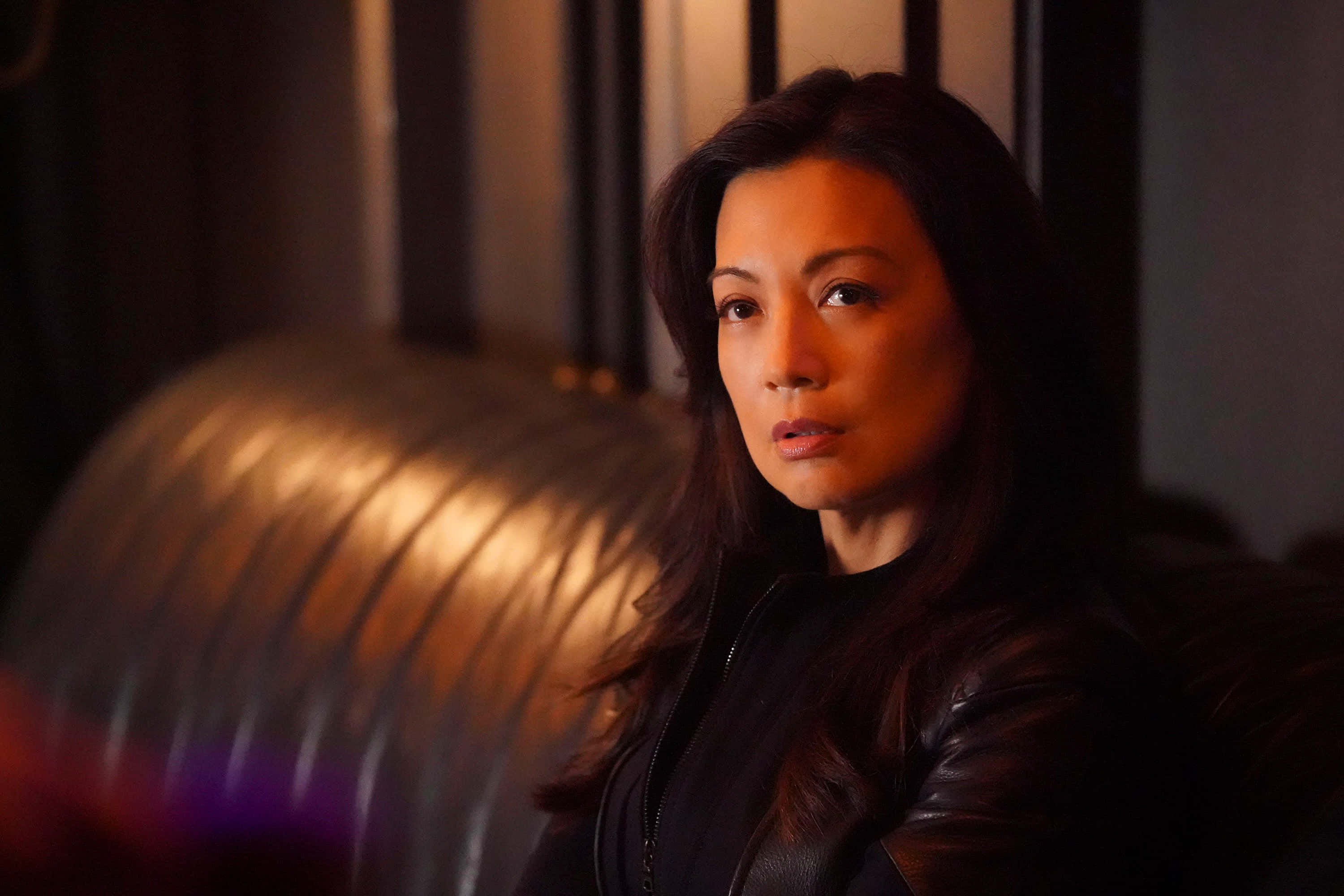 Ming Na Wen 桌布 壁紙 — 一位身穿黑色皮褸的女性，在昏暗的暖光中坐著，表情嚴肅。
