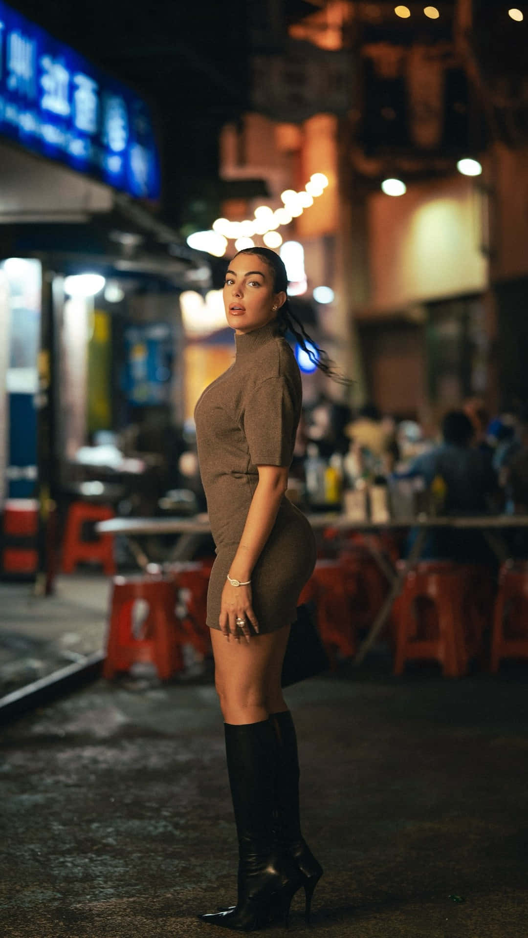  壁紙 — 一名身穿棕色連衣裙和黑色靴子的女性在夜晚霓虹燈閃爍的城市街道上自信地站立。
