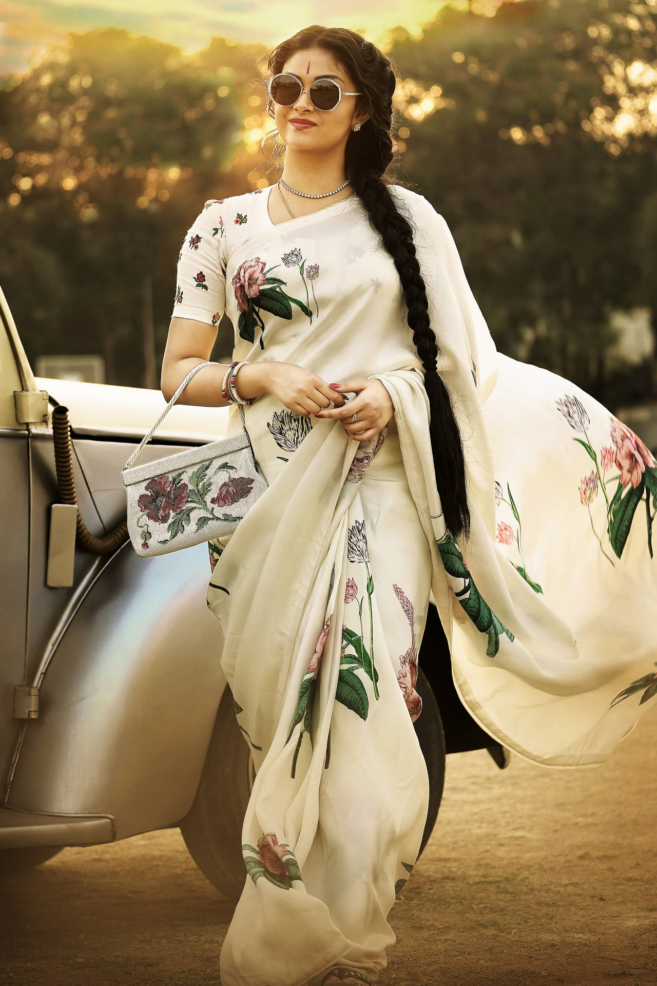 身穿花卉白色紗麗和太陽眼鏡的女性在金色夕陽下站在復古車旁。