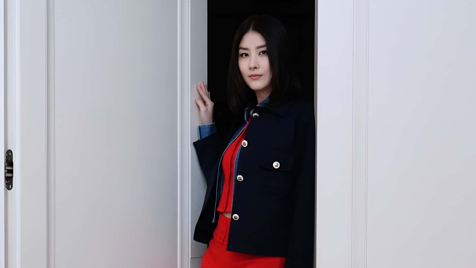  壁紙 — 一位身穿海軍藍外套和紅色服裝的女性，帶著平靜的表情從白色門後探出頭來。
