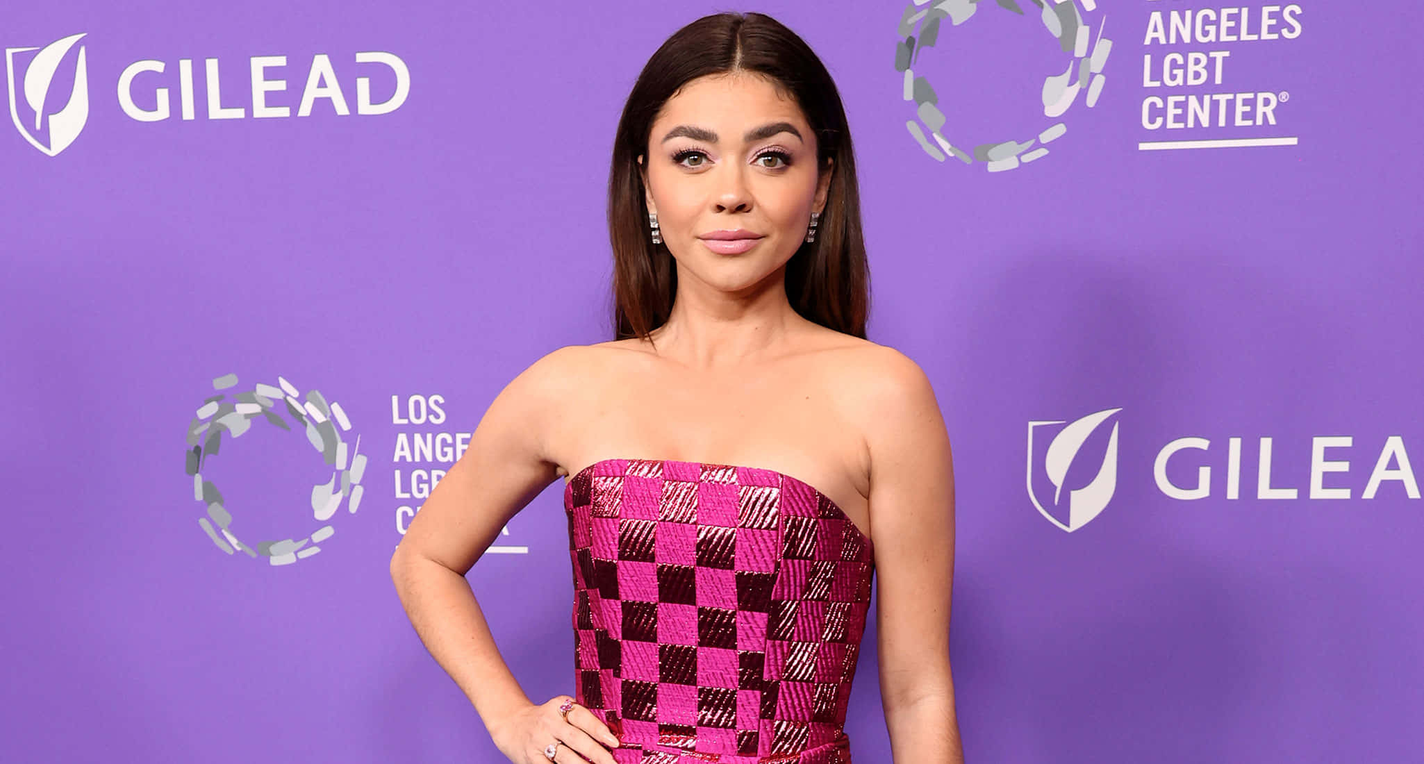 Sarah Hyland 桌布 壁紙 — 一位穿著鮮豔粉紅和黑色格紋無肩帶連衣裙的女性，在帶有標誌的紫色活動背景前自信地擺姿勢。