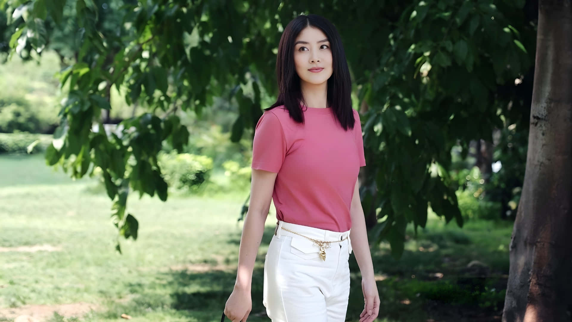  壁紙 — 一位身穿粉紅色上衣和白色褲子的女士，站在陽光普照的公園裡，樹葉茂密的樹下，光線柔和自然。