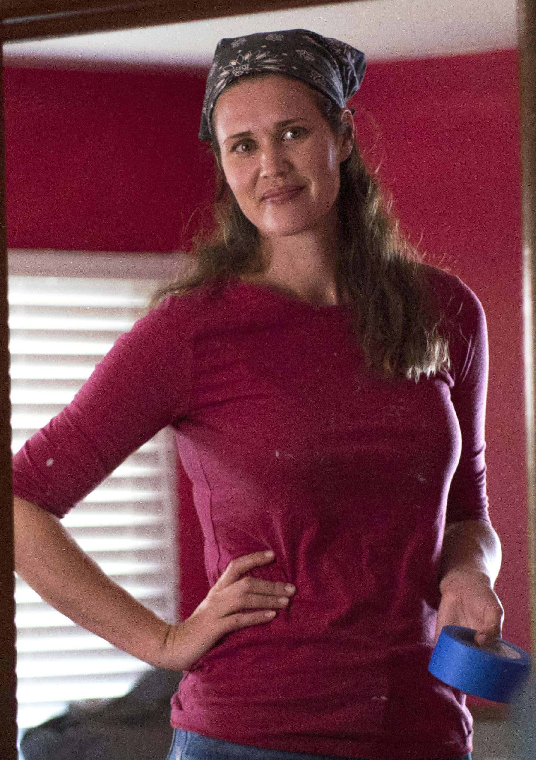 Sarah Lancaster 桌布 壁紙 — 一位身穿紅色襯衫和頭巾的女性，站在紅色牆壁和白色百葉窗的房間裡，手持藍色油漆膠帶。