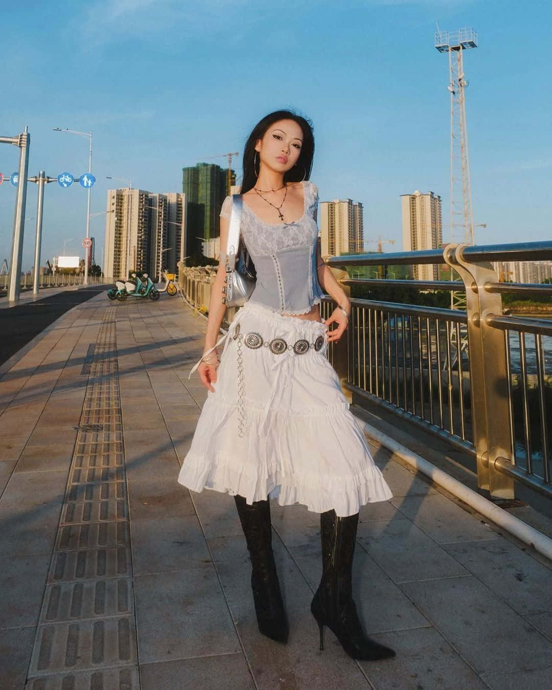  壁紙 — 一位身穿白色荷葉邊裙和黑色靴子的女性，在現代城市橋樑上自信地擺姿勢，背景是黃金時段的摩天大樓。