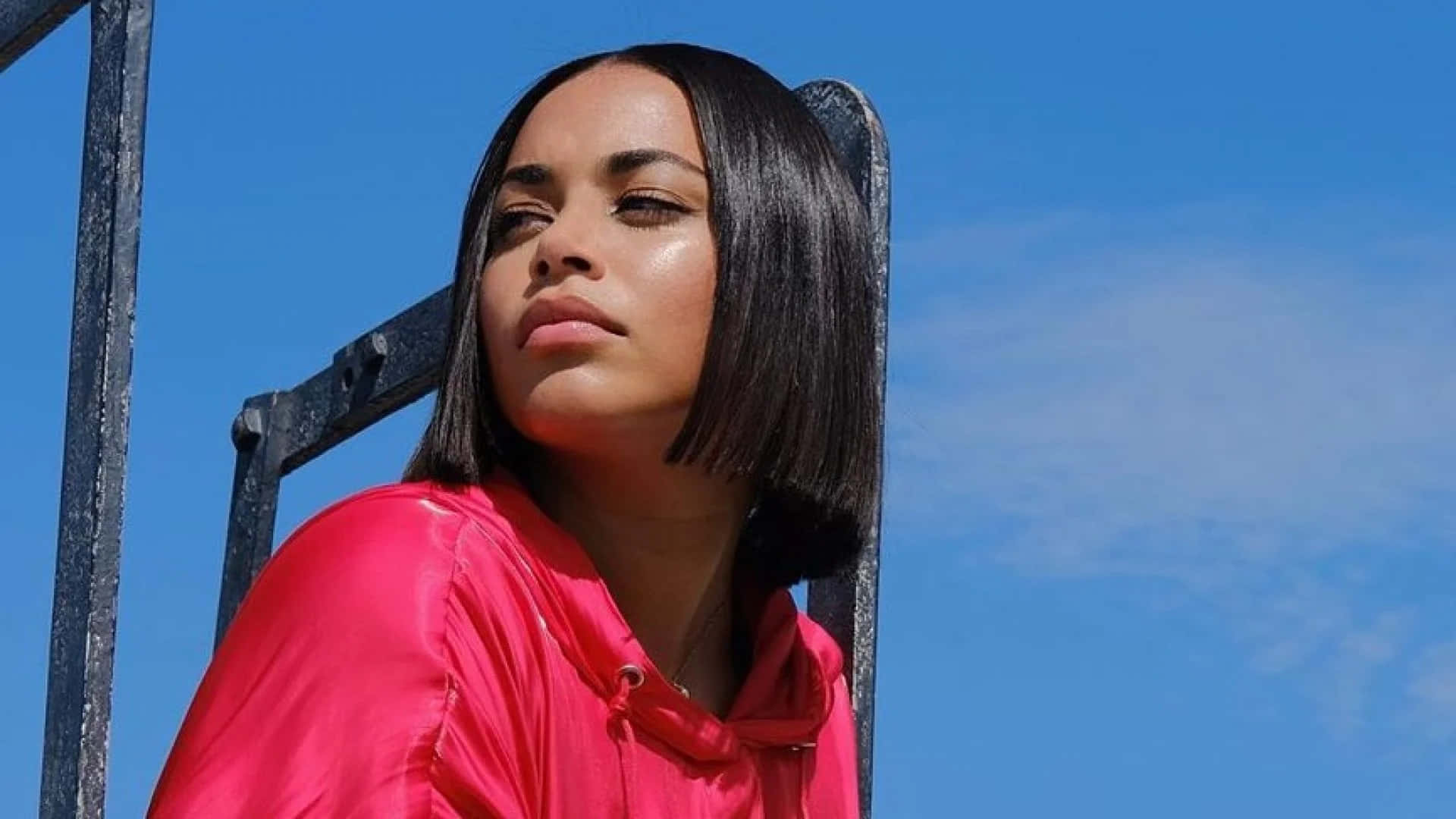Lauren London 桌布 壁紙 — 一位短鮑伯頭髮的女性穿著亮粉色連帽衫，靠在金屬結構上，背景是晴朗的藍天。