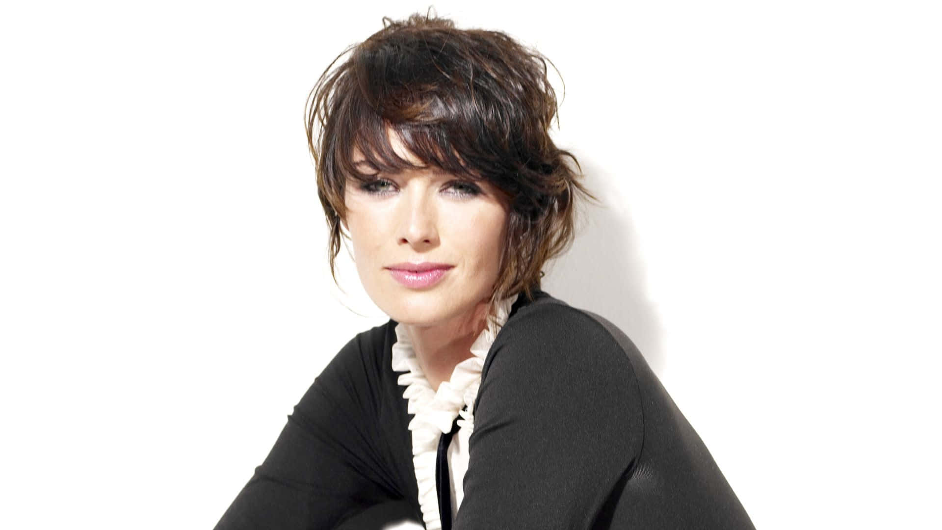 Lena Headey 桌布 壁紙 — 一位短黑髮女性，身穿黑色上衣配白色荷葉邊領，在純白背景前擺姿勢。