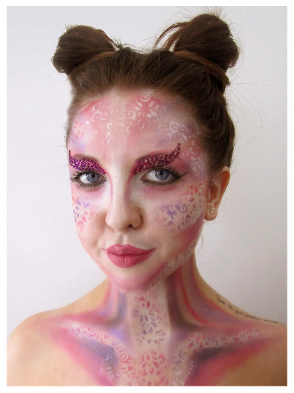 Fantasy Makeup 桌布 壁紙 — 一位女士臉上和頸部帶有精緻的粉紅色和紫色閃粉化妝圖案，頭髮梳成兩個髮髻，背景為純白色。