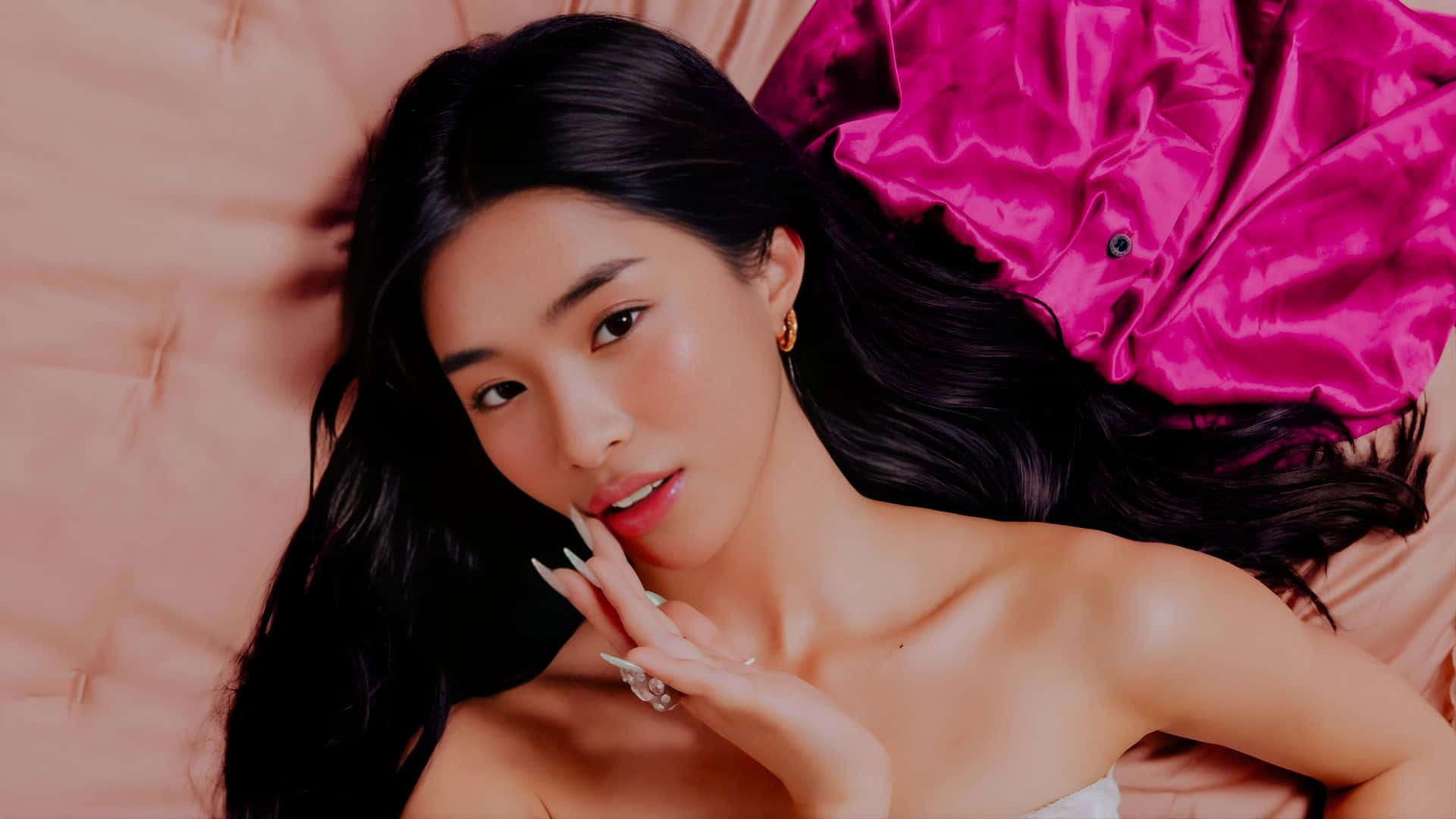  壁紙 — 一位長黑髮的女性躺在粉紅色簇絨床上，溫柔地凝視，旁邊放著一件鮮豔的洋紅色緞面衣物。