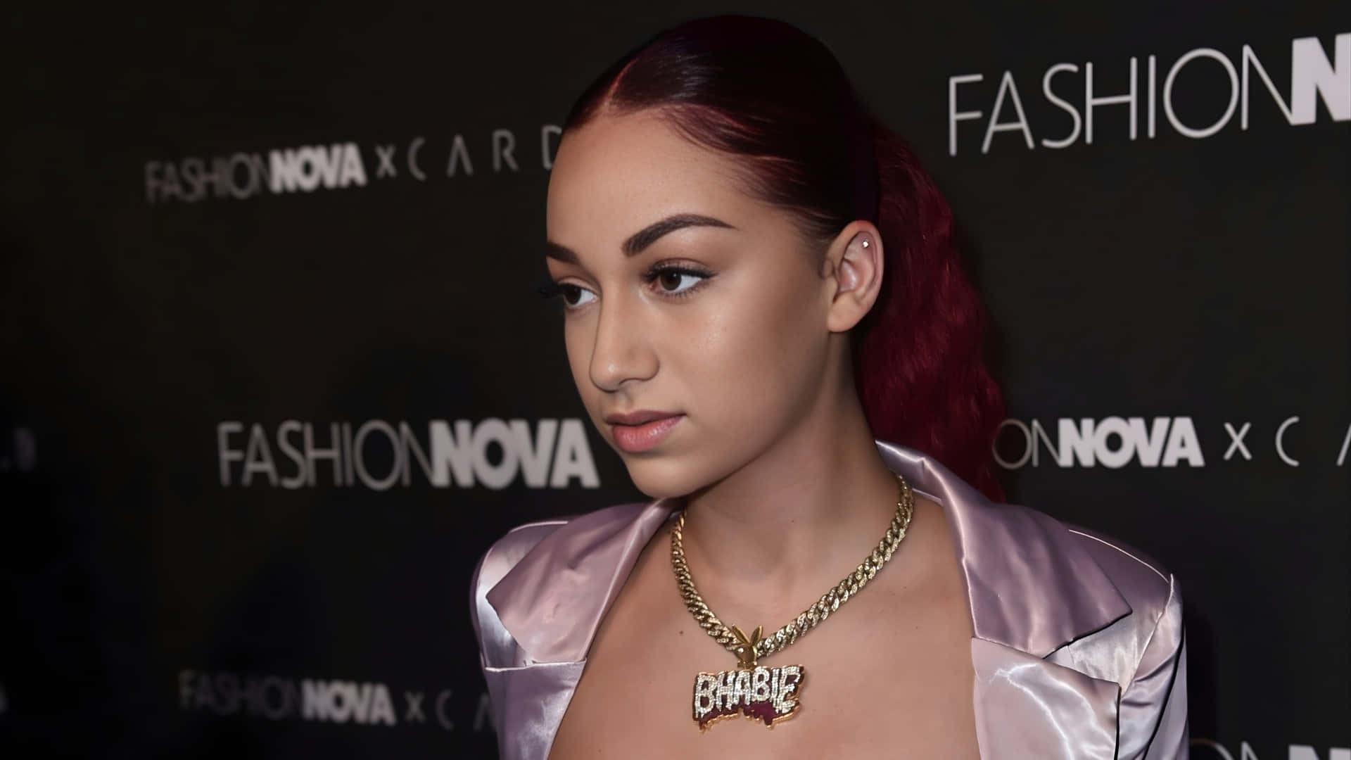  壁紙 — 一位紅髮馬尾辮的女性在Fashion Nova活動中佩戴著帶有'BHABIE'吊墜的金鏈項鍊和粉紅色緞面外套。