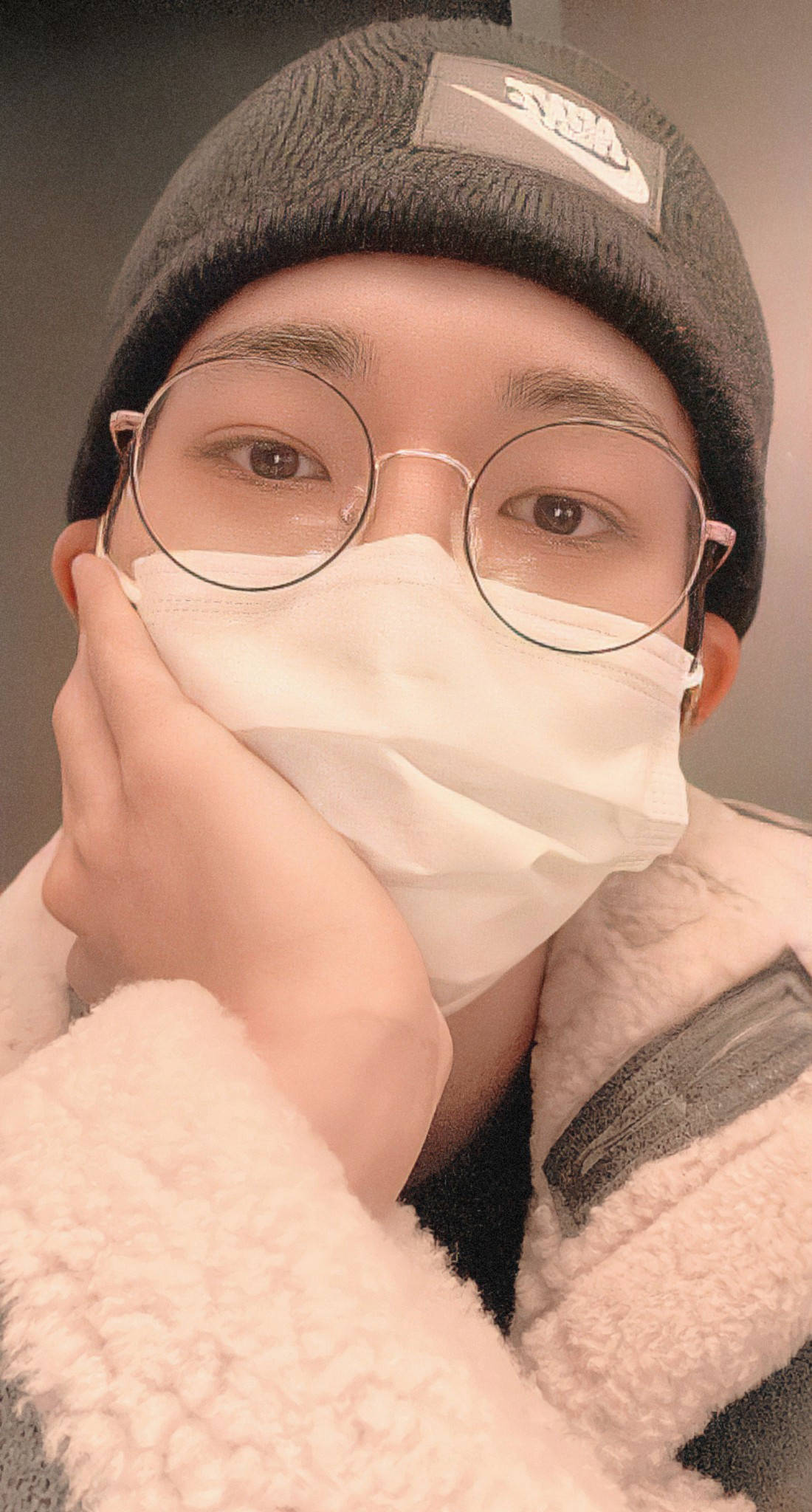 Wonwoo 戴口罩 壁紙 — 一位戴著圓框眼鏡和白色口罩的年輕人，托著下巴，身穿灰色冷帽和絨毛外套。