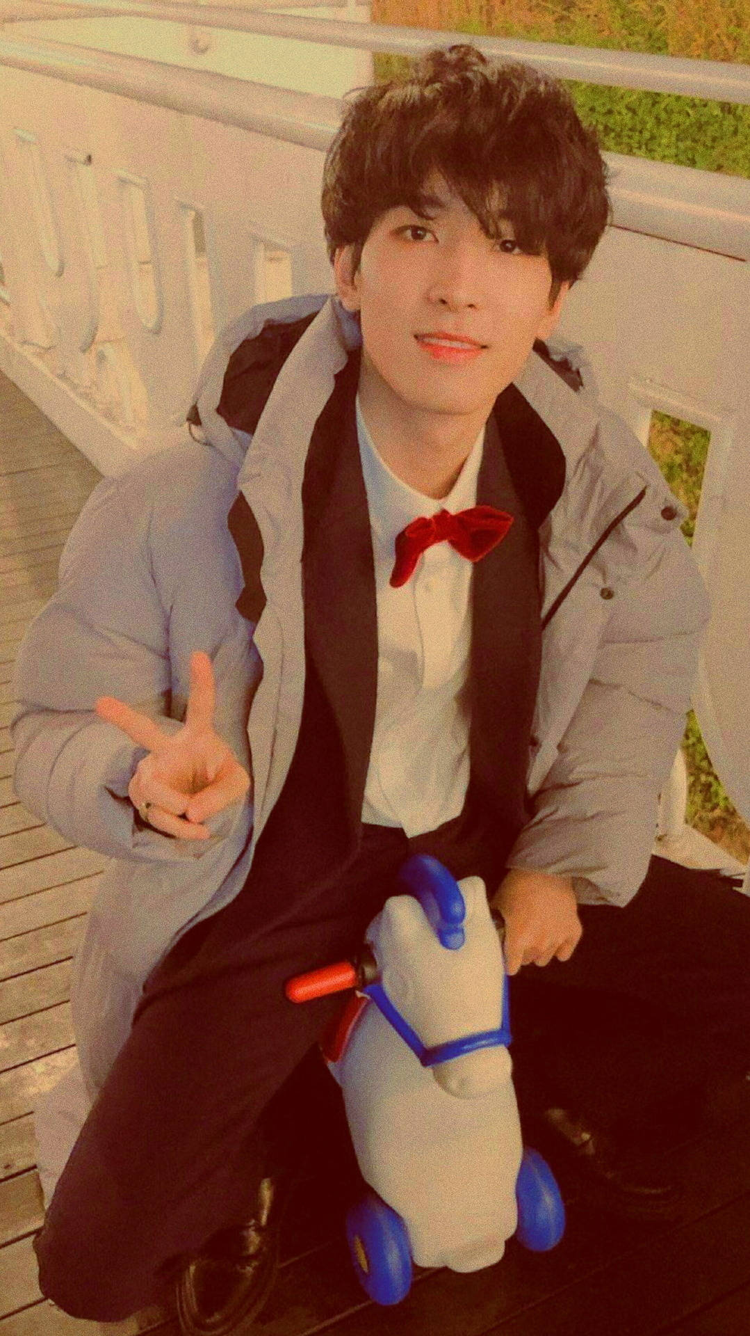 Wonwoo 騎搖搖馬 壁紙 — 一位身穿淺灰色羽絨外套和紅色領結的年輕男子微笑著坐在木甲板上，旁邊是一隻白色和藍色的玩具馬。