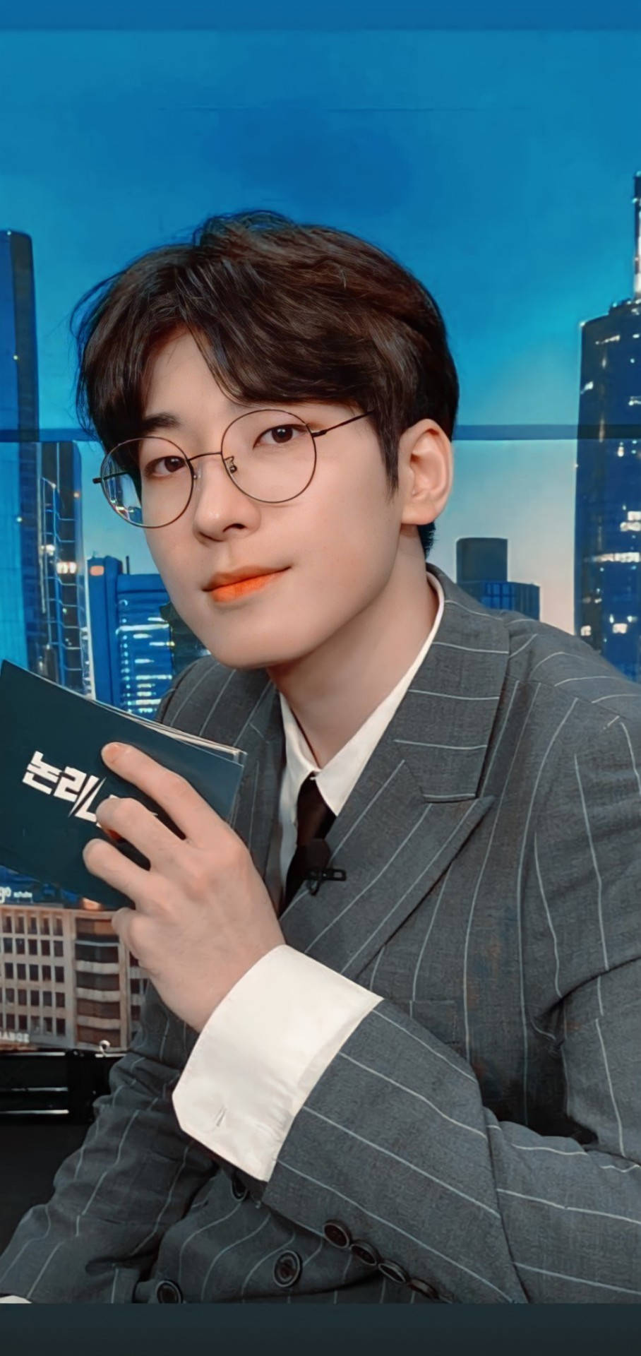 Wonwoo《Going Seventeen》 壁紙 — 一位身穿細條紋西裝、戴眼鏡的年輕男子，以充滿活力的藍色城市景觀為背景，手持一本書。