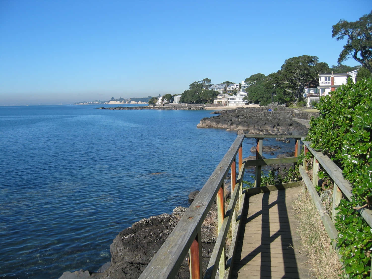 Seaside 桌布 壁紙 — 一條木製步道蜿蜒在岩石海岸線上，平靜的藍色海水和白色房屋隱藏在綠樹之間，在晴朗的天空下。