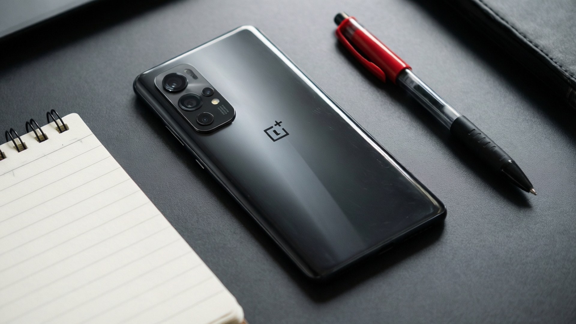 黑色OnePlus手機、紅色筆和橫線筆記本在深灰色桌上
