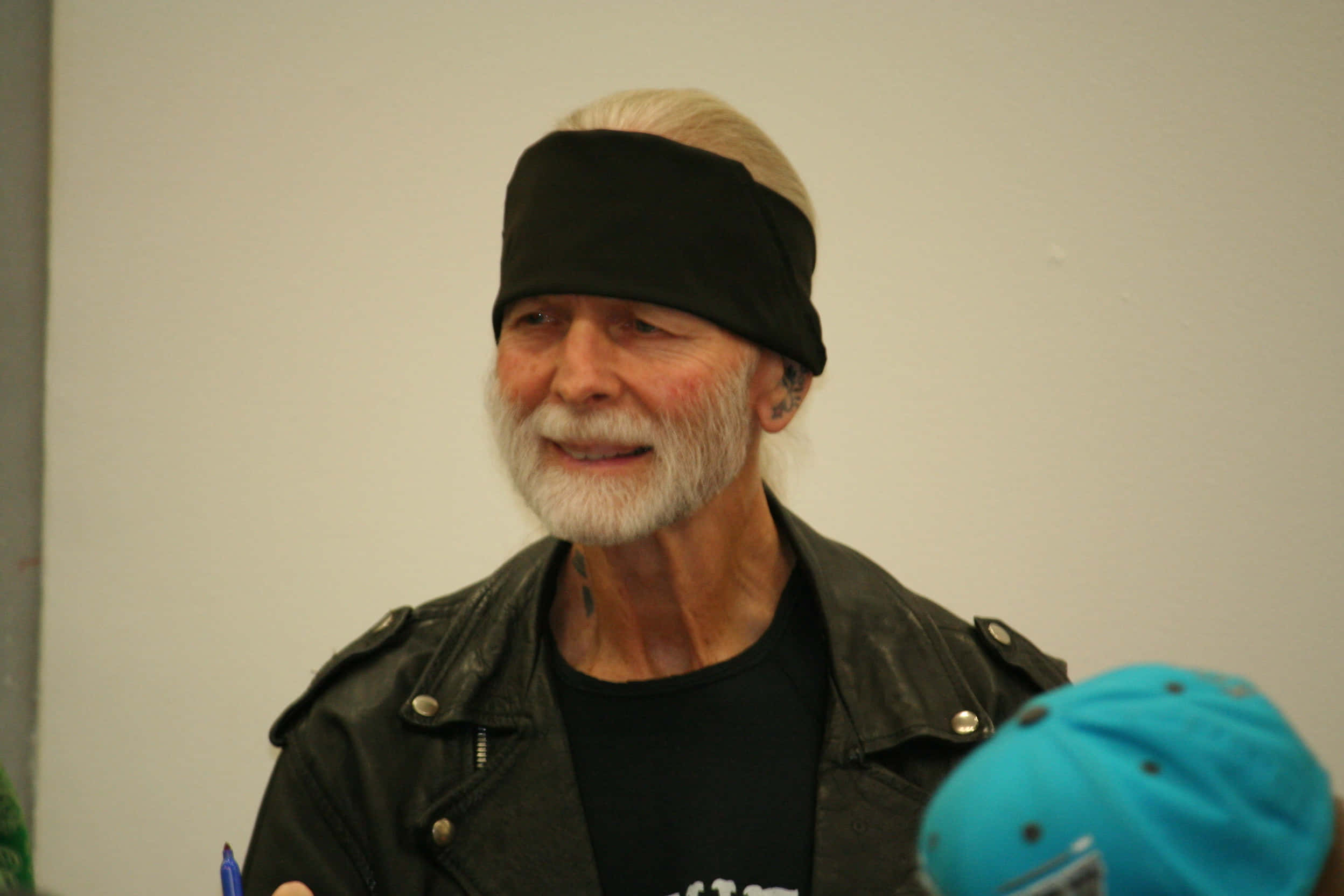 吉米·瓦利安特 WrestleCade 2012 壁紙 — 一位留著白鬍子、戴黑色頭帶的長者，在室內穿著皮褸微笑。